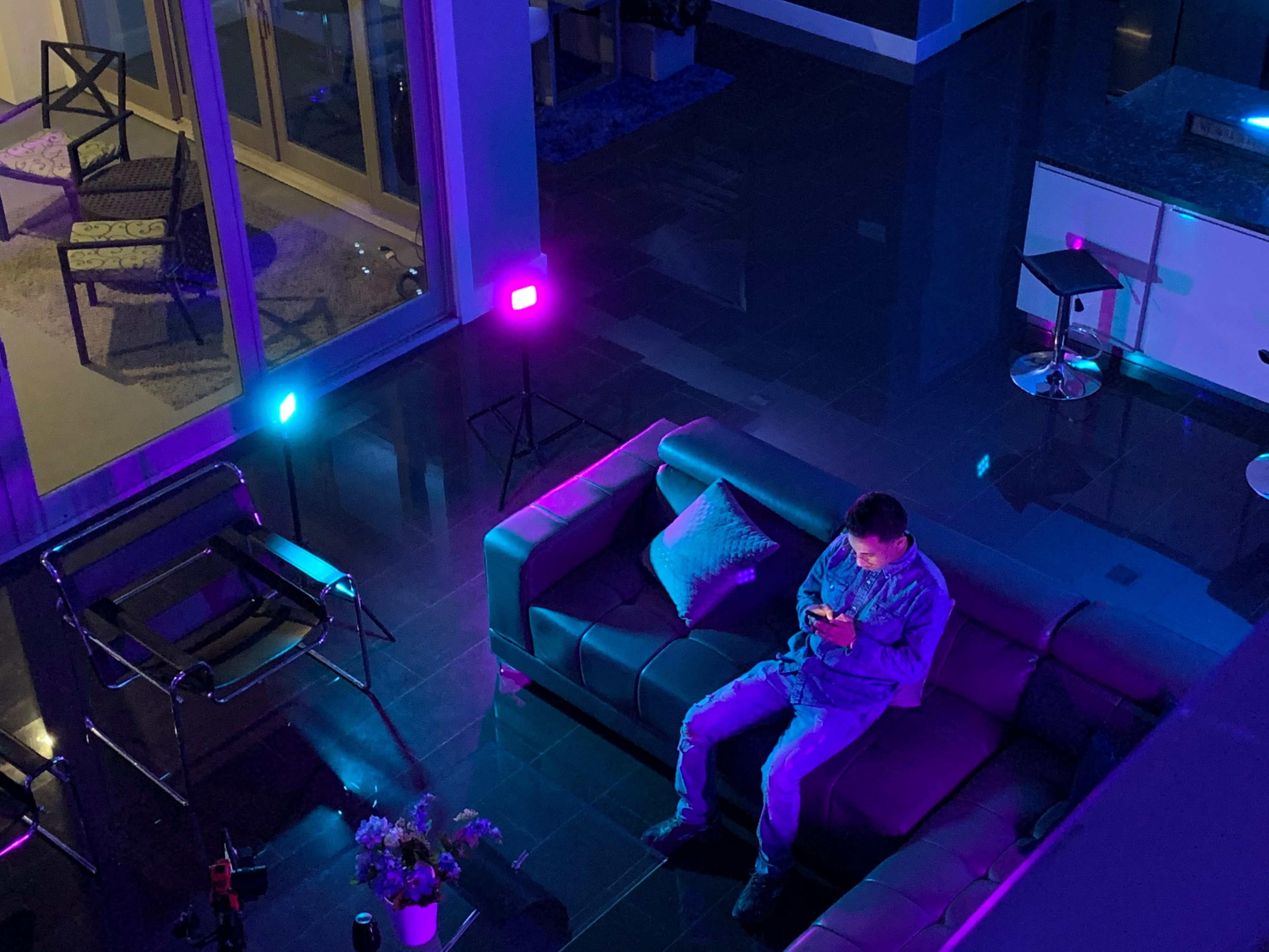 A person sits on a dark-gray sofa in a dimly lit room, looking at a phone while colorful lights illuminate the space.