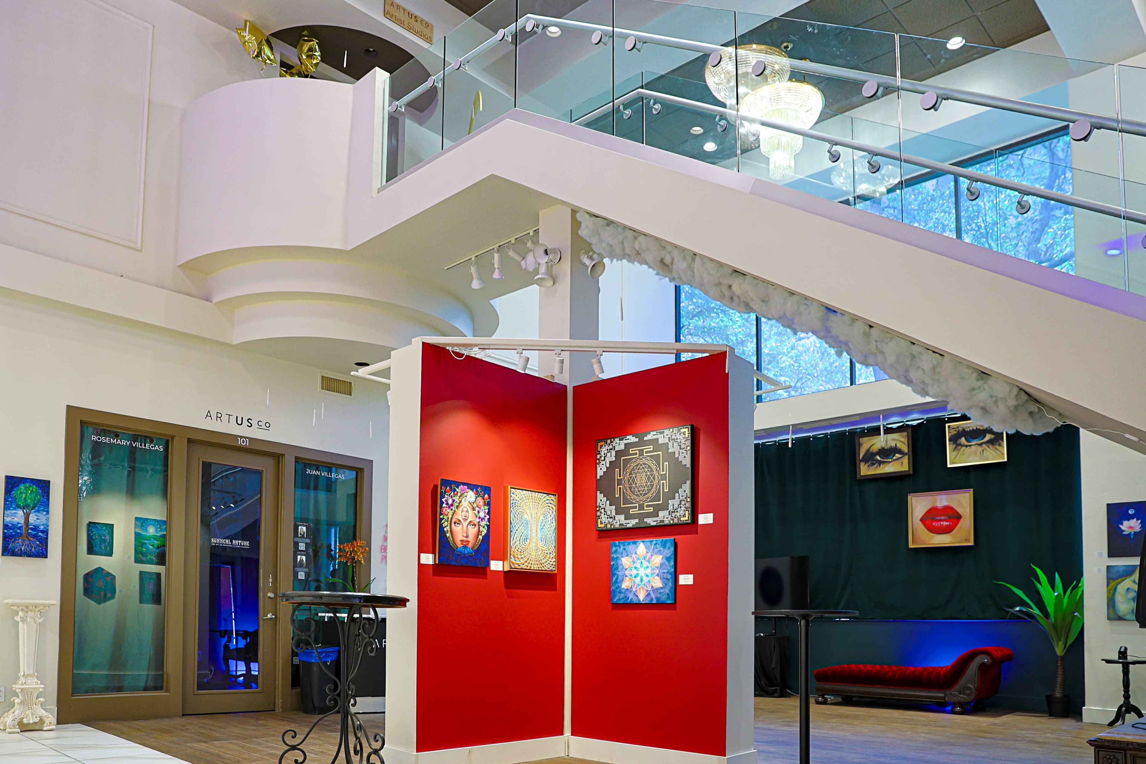 An art gallery featuring a red partition with framed artworks, a staircase, and various decorative elements in a bright indoor space.
