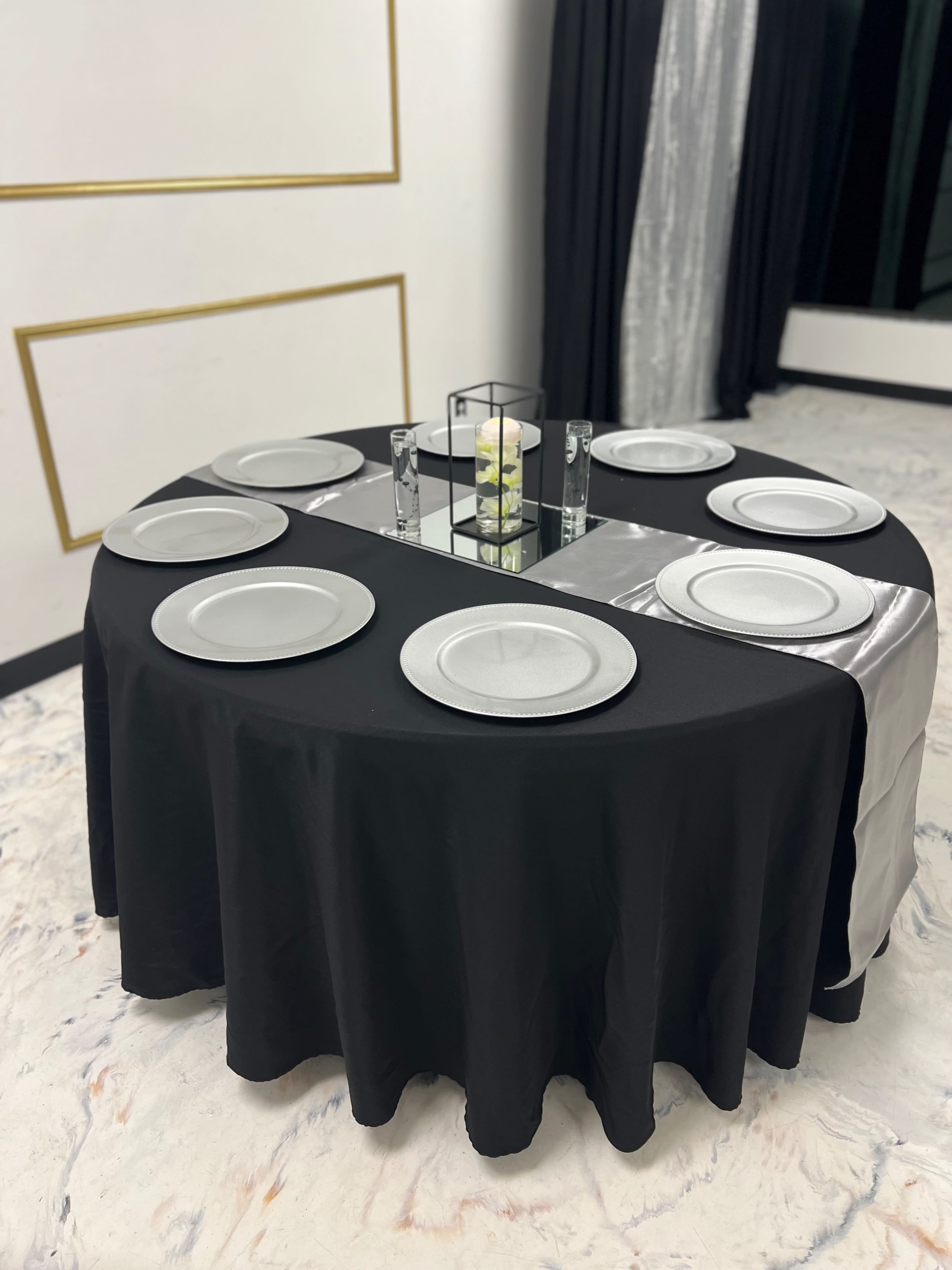 A round table covered with a black and silver tablecloth is set with six silver dinner plates and a small centerpiece.