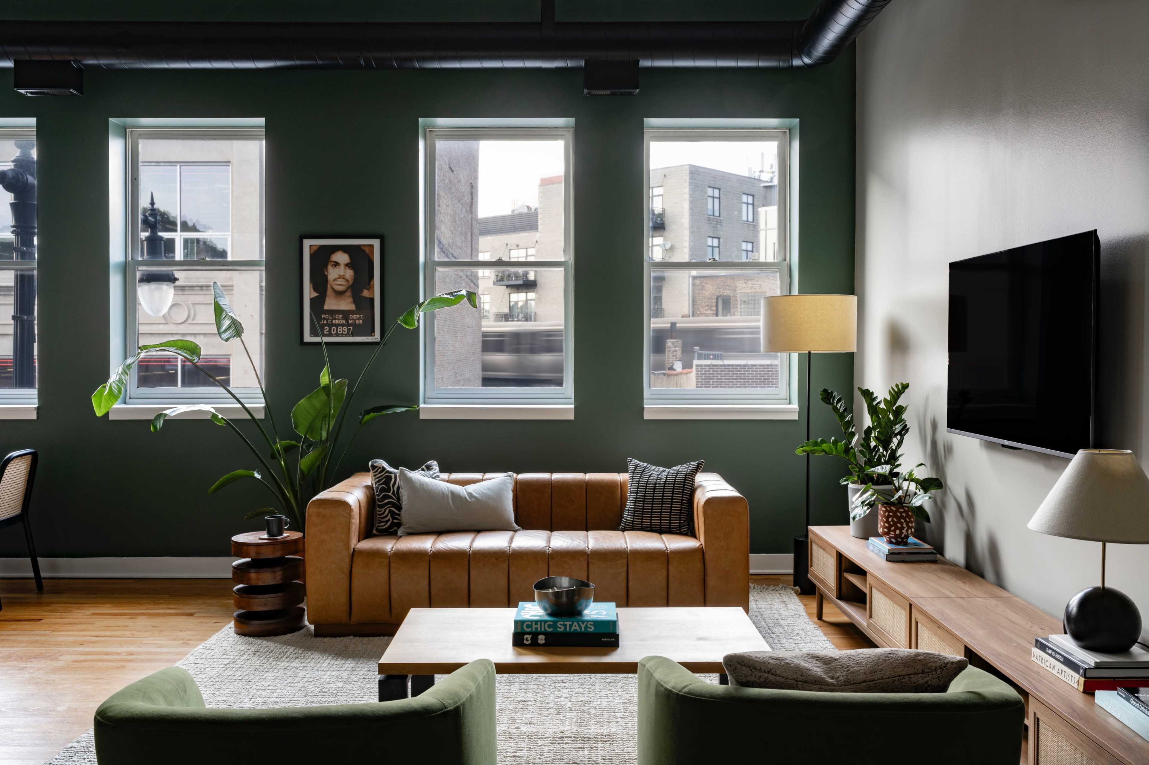 The image depicts a modern living room featuring a brown leather sofa, a green accent wall, large windows, and a television mounted on the wall.