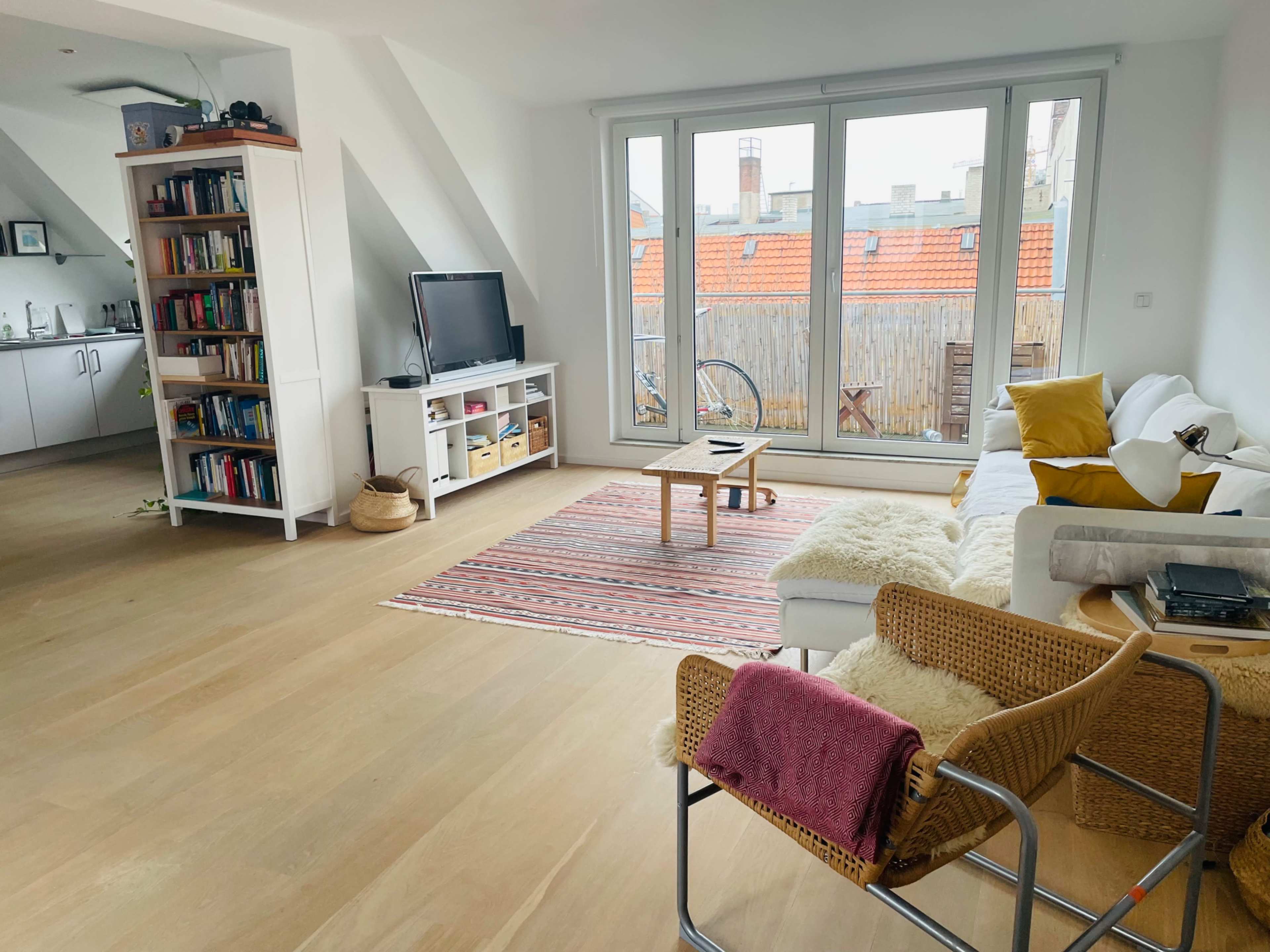 A bright living room features a white sofa, a coffee table on a patterned rug, a bookshelf filled with books, and large windows overlooking a courtyard.