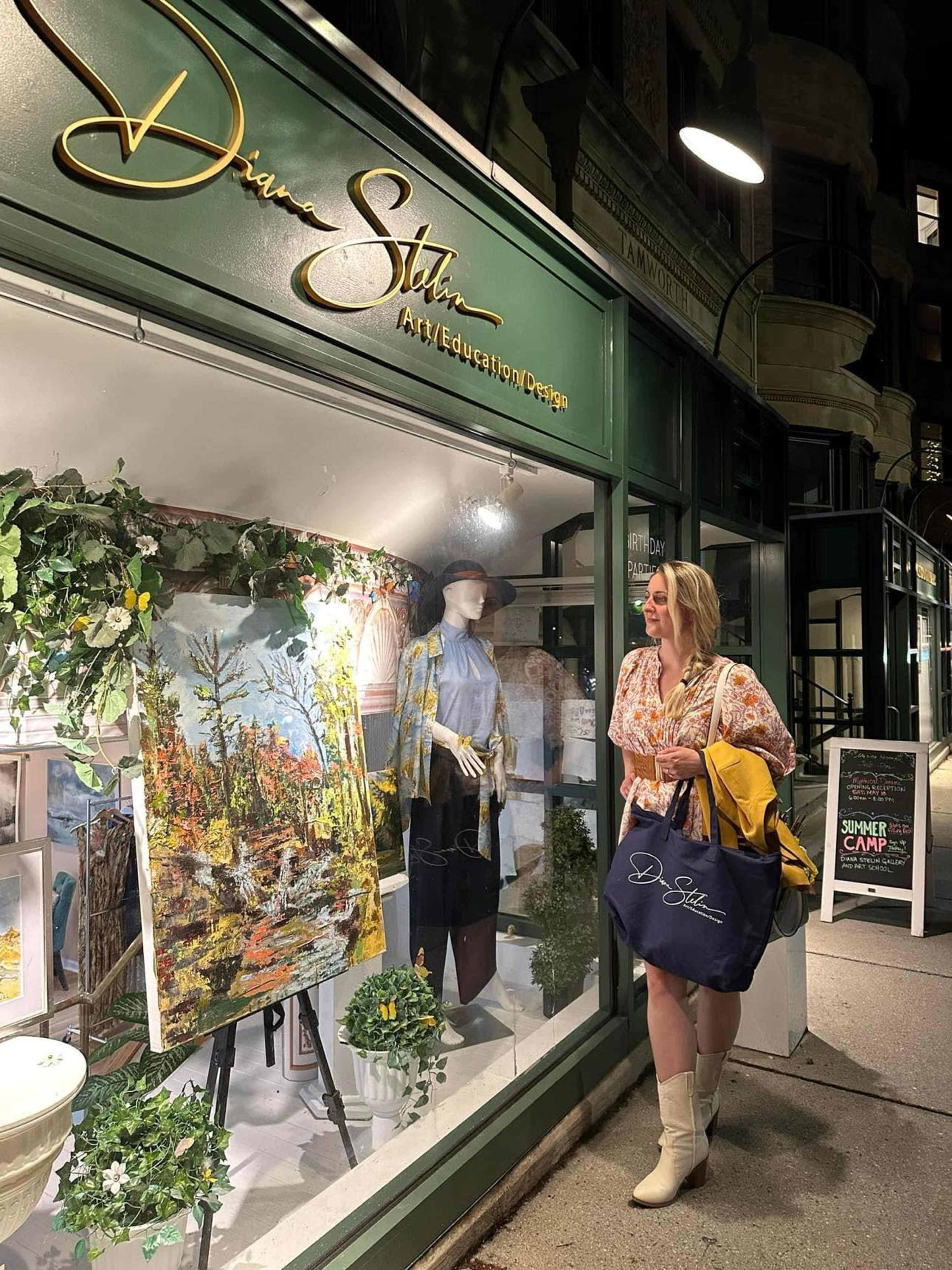 A woman wearing a floral blouse and boots stands in front of an art gallery window displaying a colorful landscape painting and a mannequin dressed in a stylish outfit.
