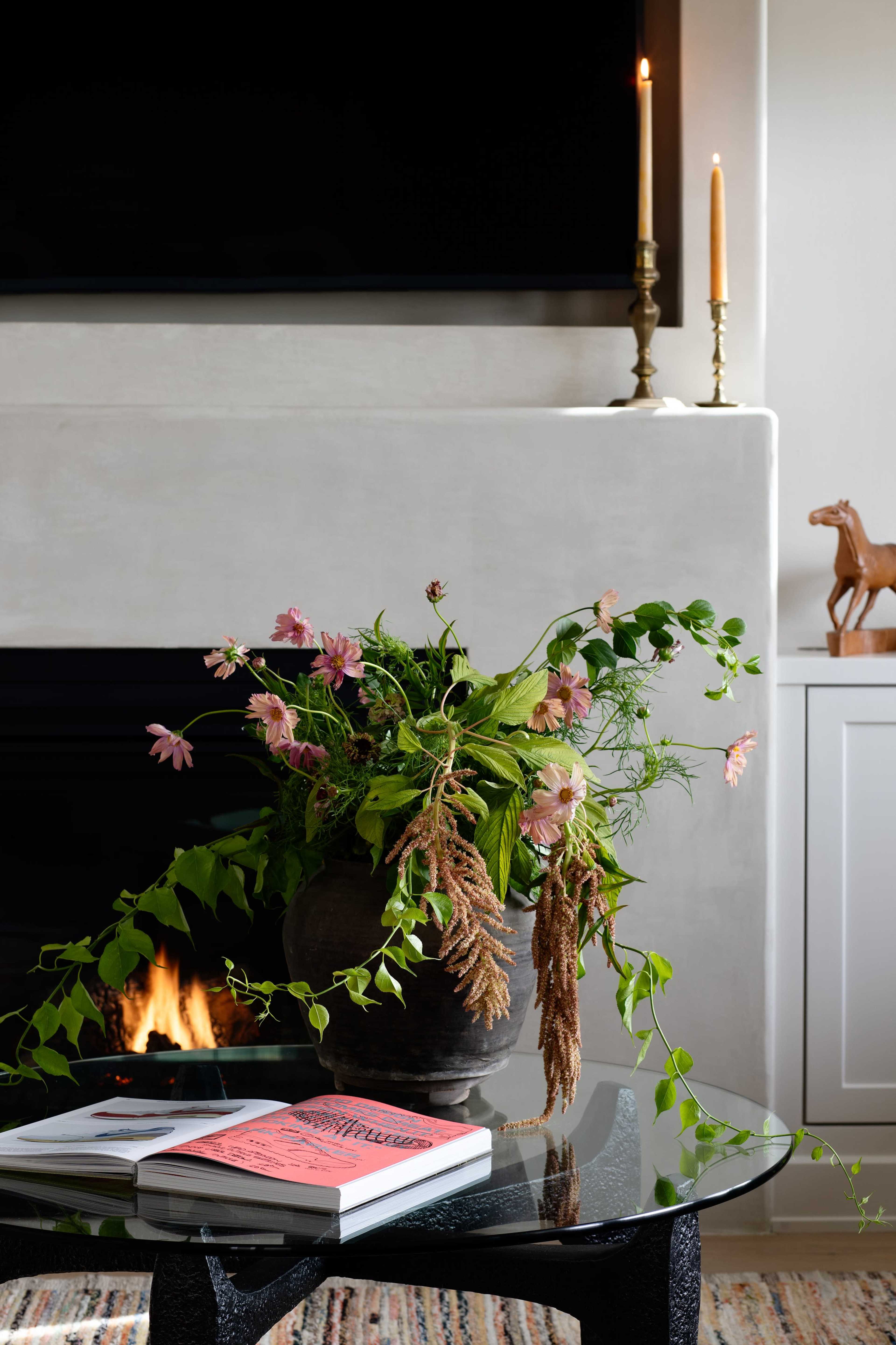 A vase of flowers is prominently displayed on a glass coffee table in a cozy living room with a lit fireplace and candles.