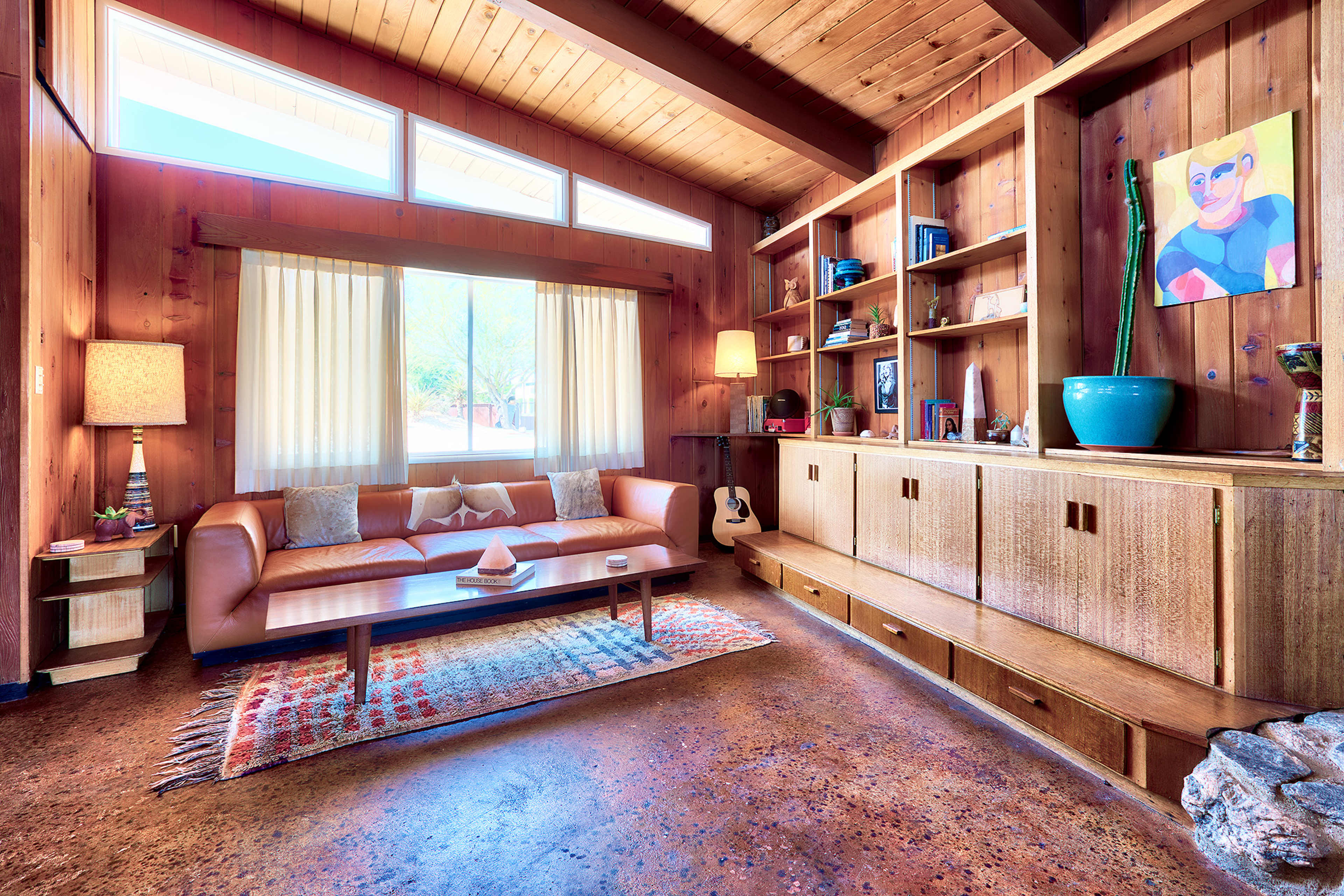 A well-lit living room features a tan sofa, a wooden coffee table, a bookshelf with decorative items, and a large window with sheer curtains.