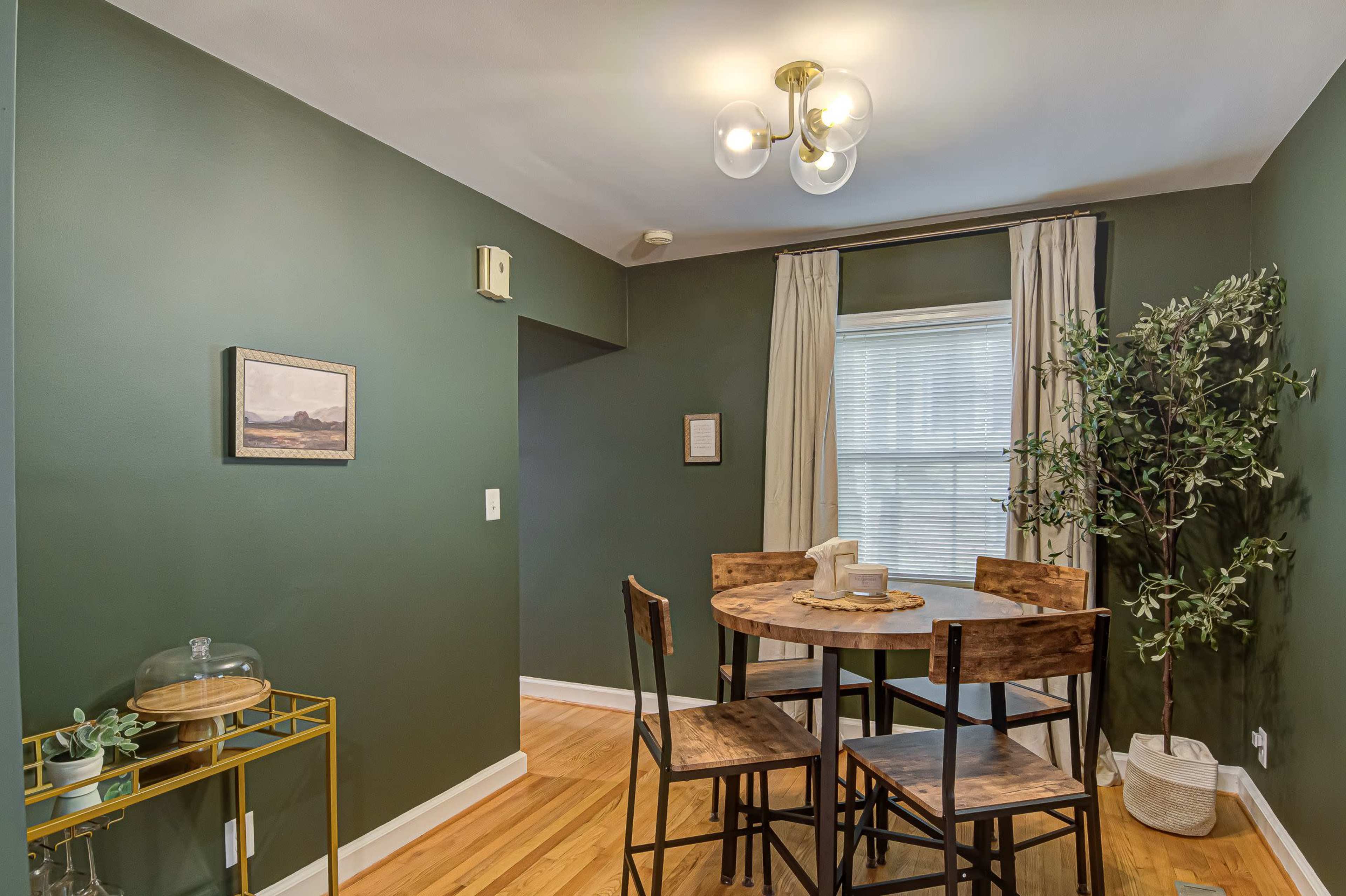 The image shows a small dining area with a circular wooden table surrounded by four chairs, a green painted wall, and a decorative plant in a white pot.