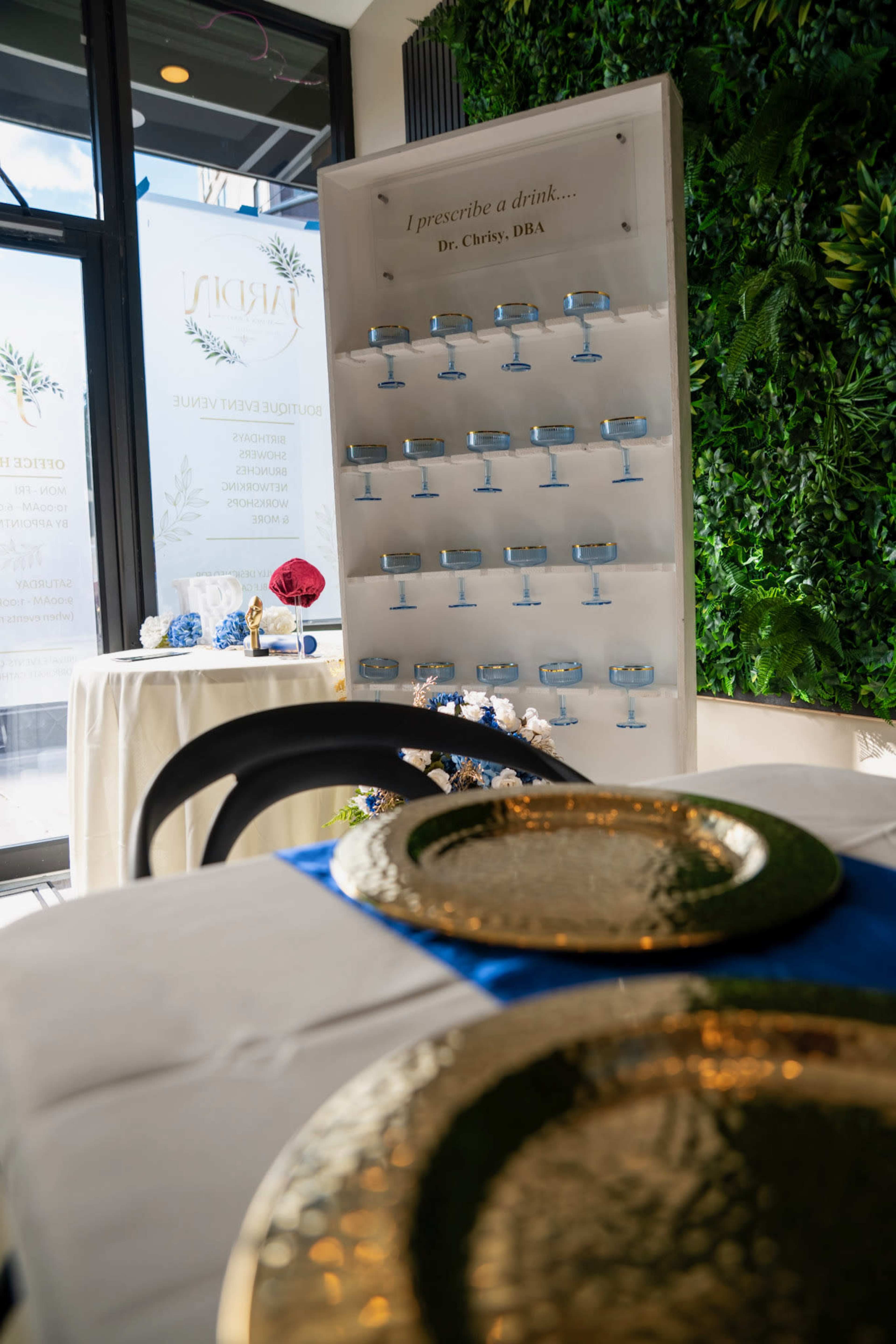 A table set with gold-rimmed plates and a blue table runner is displayed in front of a wall featuring drink prescriptions in a decorated indoor space.
