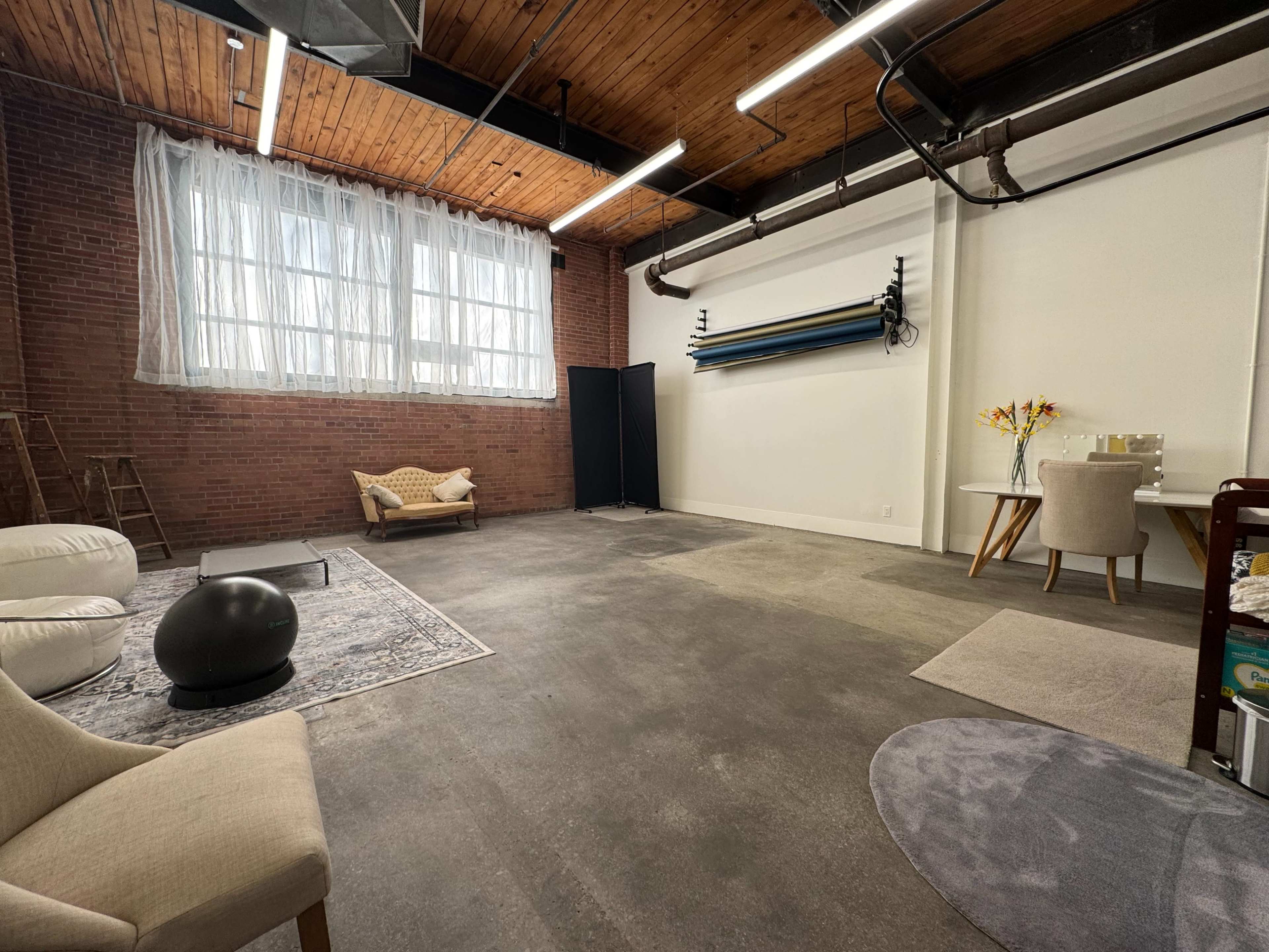 The image shows a spacious, industrial-style interior with hardwood ceilings, brick walls, and a mix of seating options, including a chaise lounge, a round table, and bean bags.