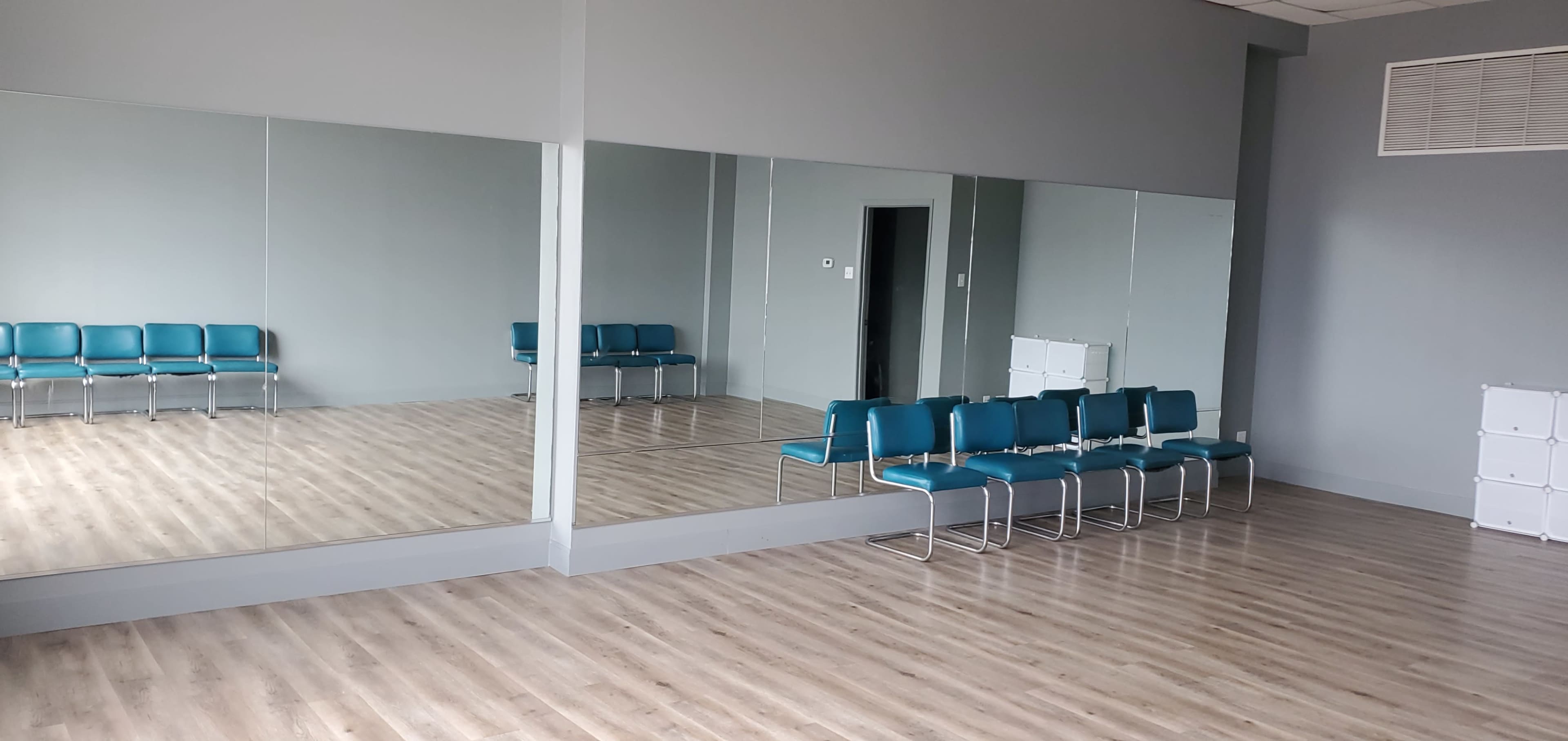 A spacious room with gray walls, wooden flooring, and large mirrors, featuring a row of teal chairs along one side.