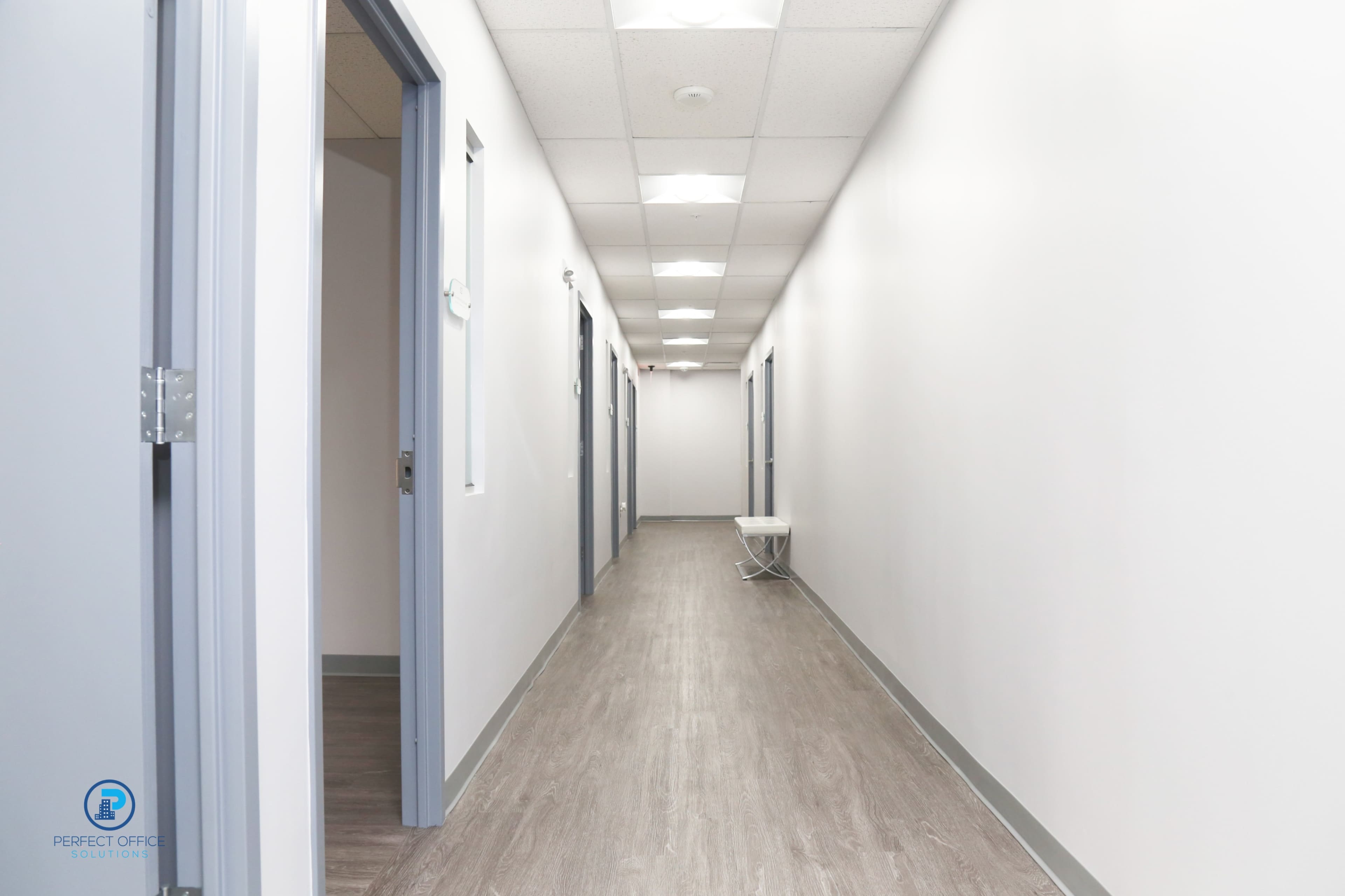 A long, well-lit corridor with multiple closed doors on either side and a folding chair positioned against one wall.