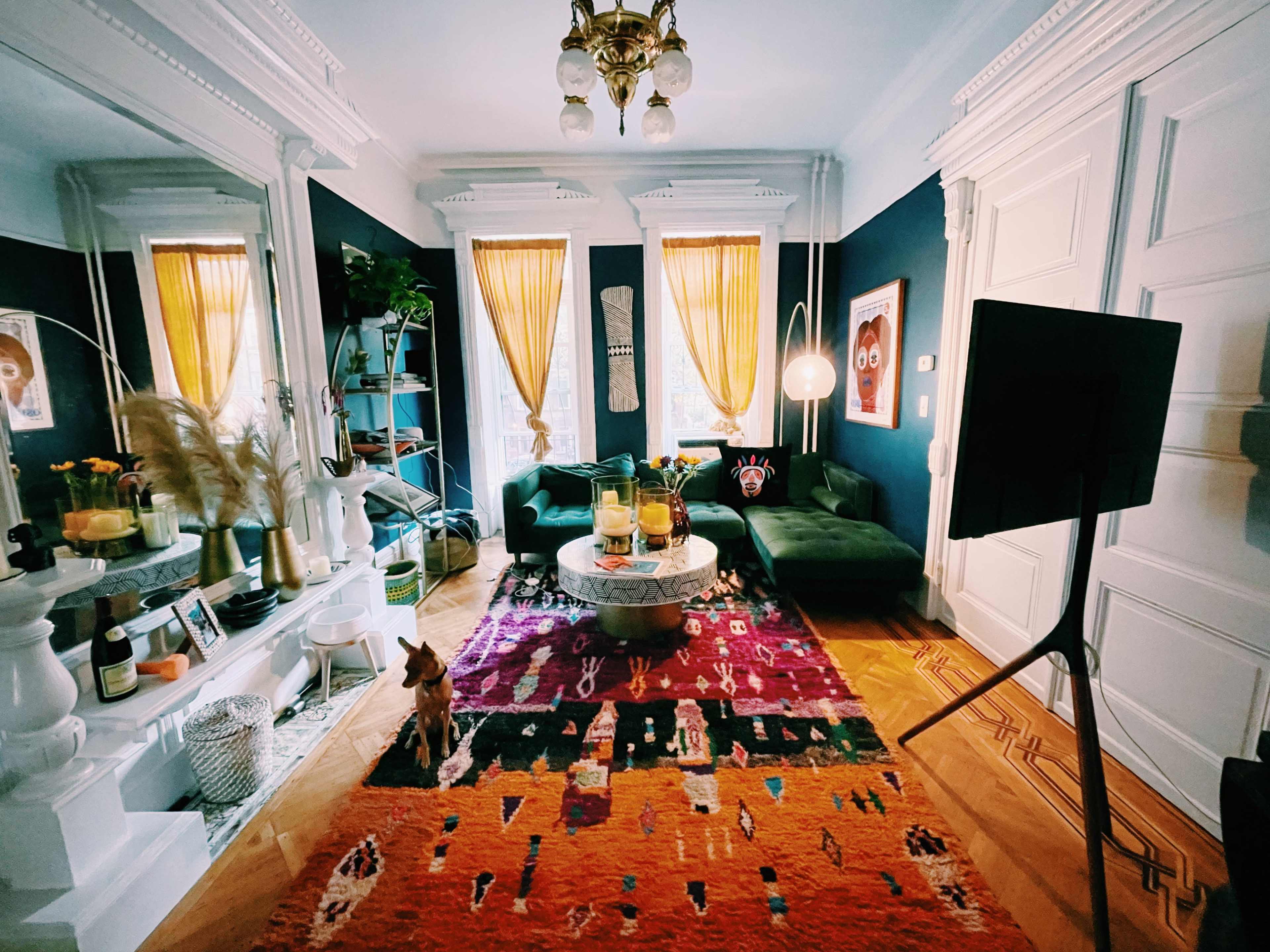 A stylish living room featuring green sofas, a round coffee table, a colorful patterned rug, and mirrored walls with large windows adorned with yellow curtains.