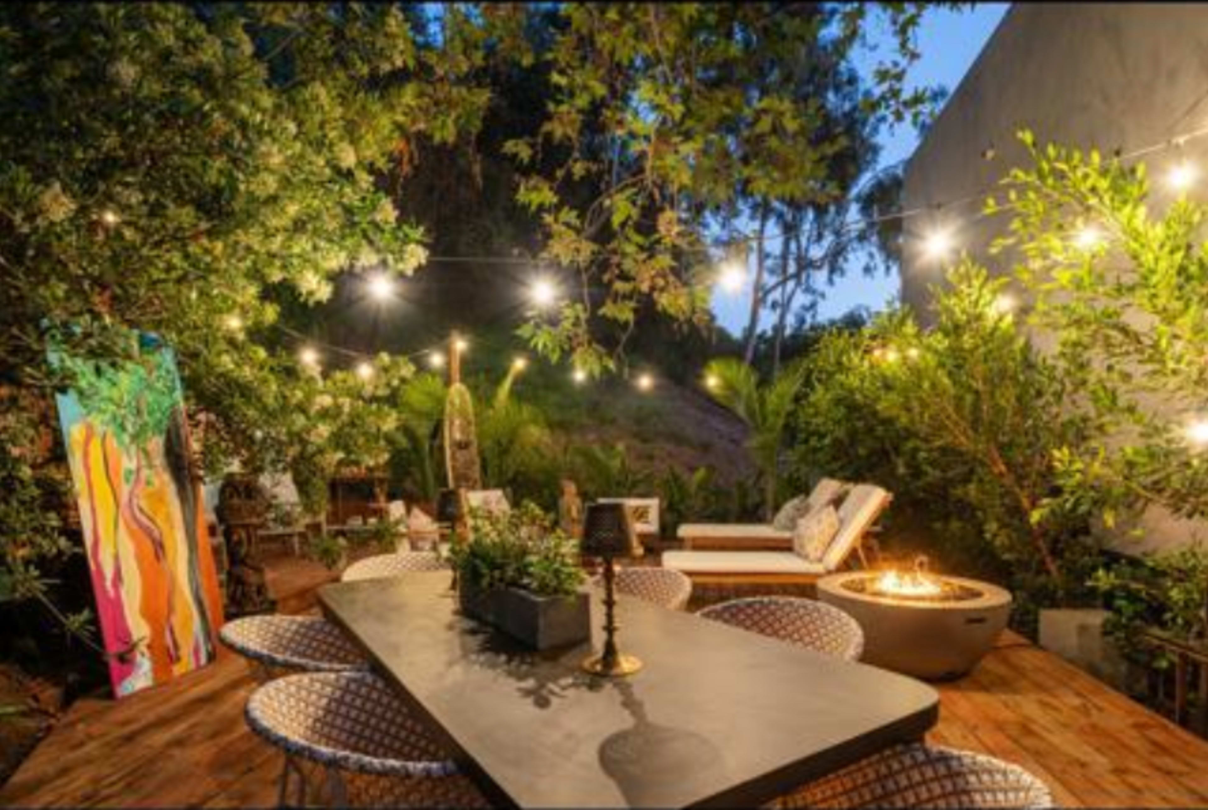 A well-lit outdoor patio features a large table, cozy seating, a fire pit, and decorative string lights amidst lush greenery.