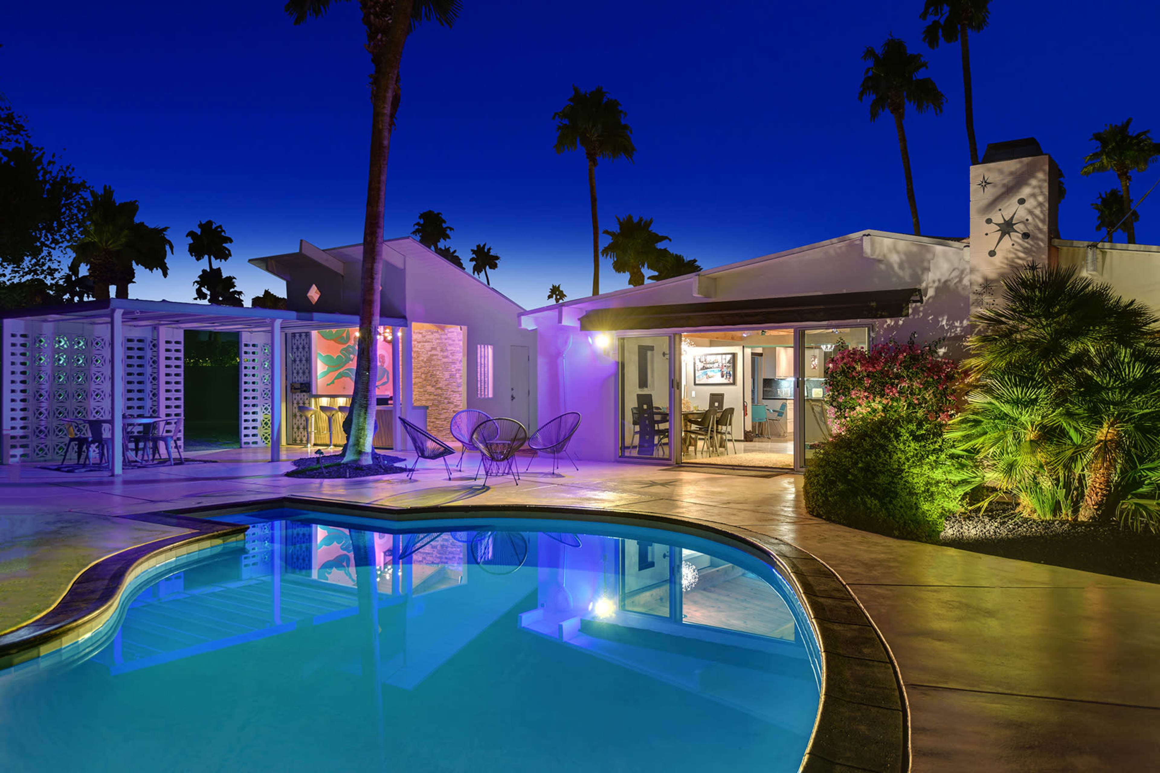 A modern house with a pool is illuminated at twilight, surrounded by palm trees and vibrant landscaping.