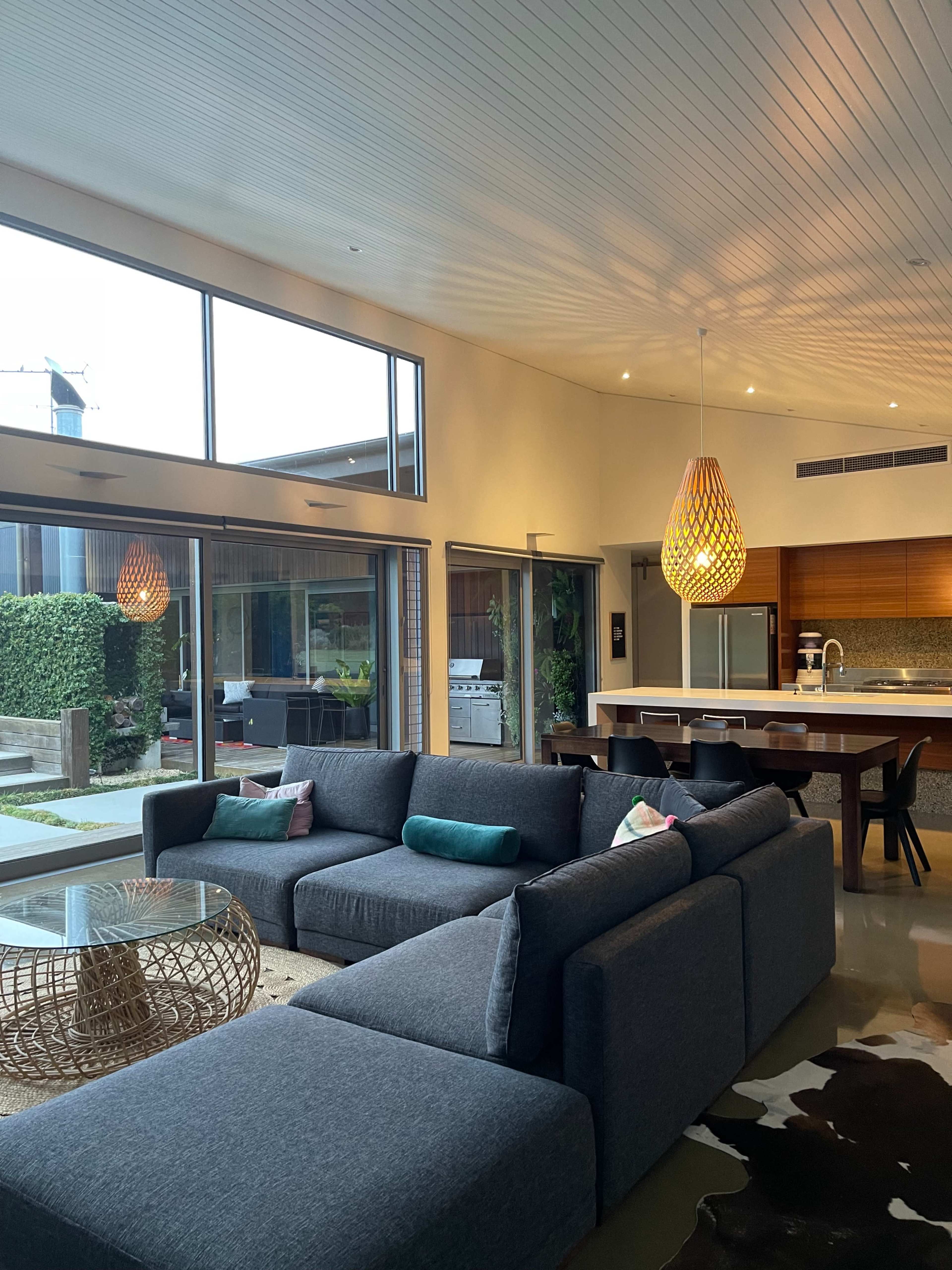 The image shows a modern living room featuring a large sectional sofa, a glass coffee table, and large windows that let in natural light.