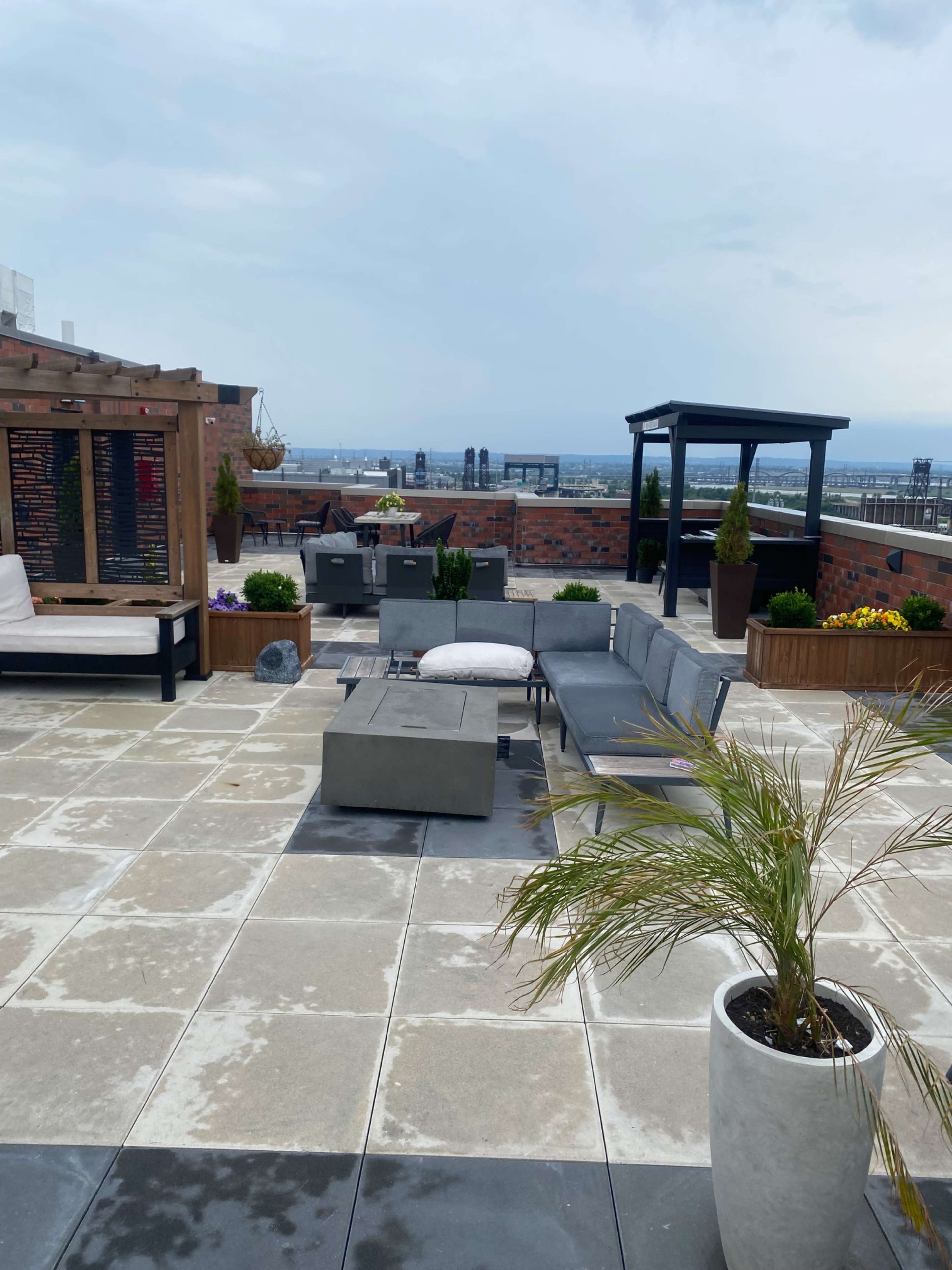 The image shows a rooftop terrace with modern seating arrangements, planters, and a view of the skyline in the background.