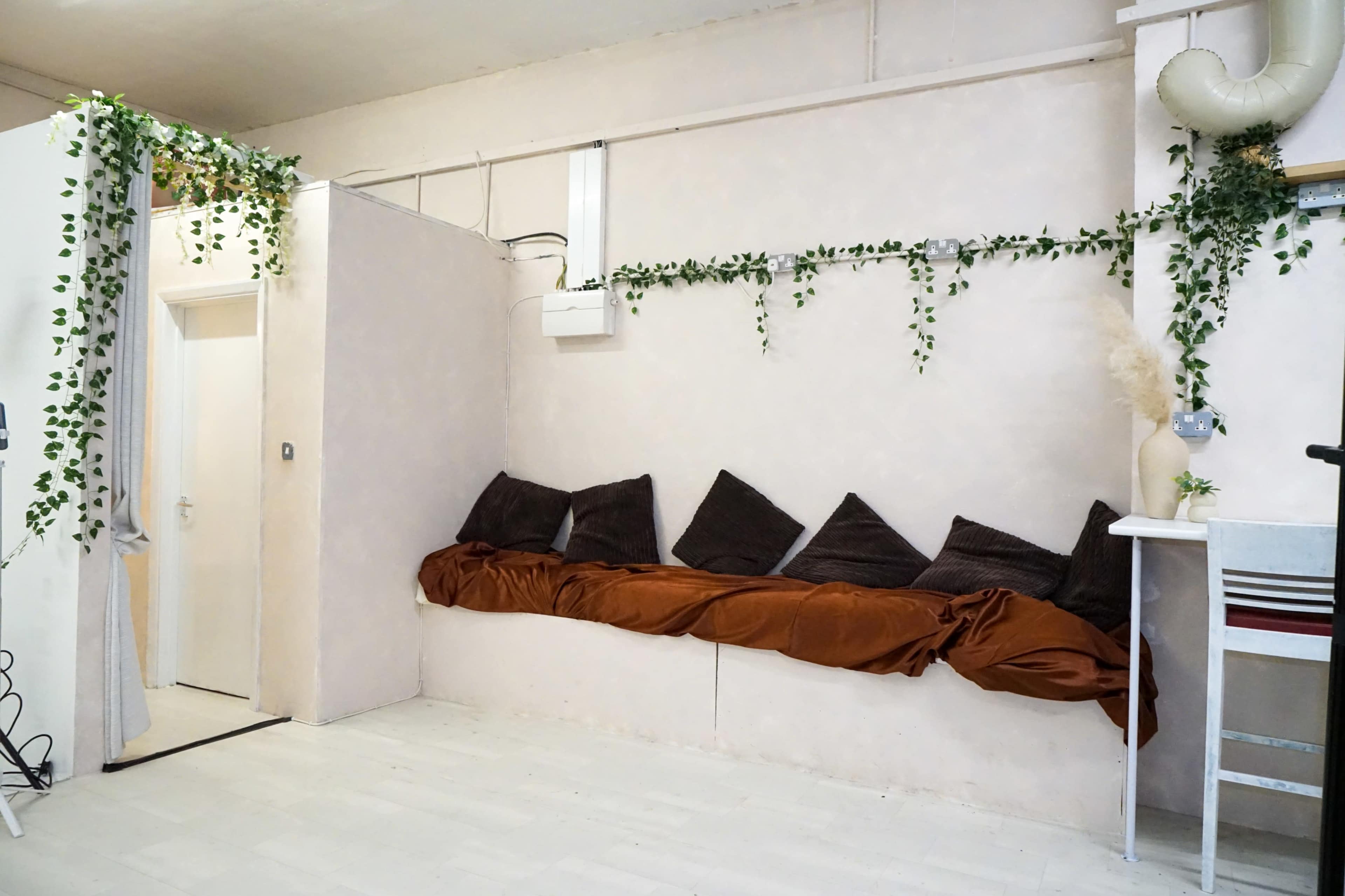 The image shows a minimalist interior with a long couch covered in a brown fabric, against a pale wall adorned with green foliage and a door leading to another room.