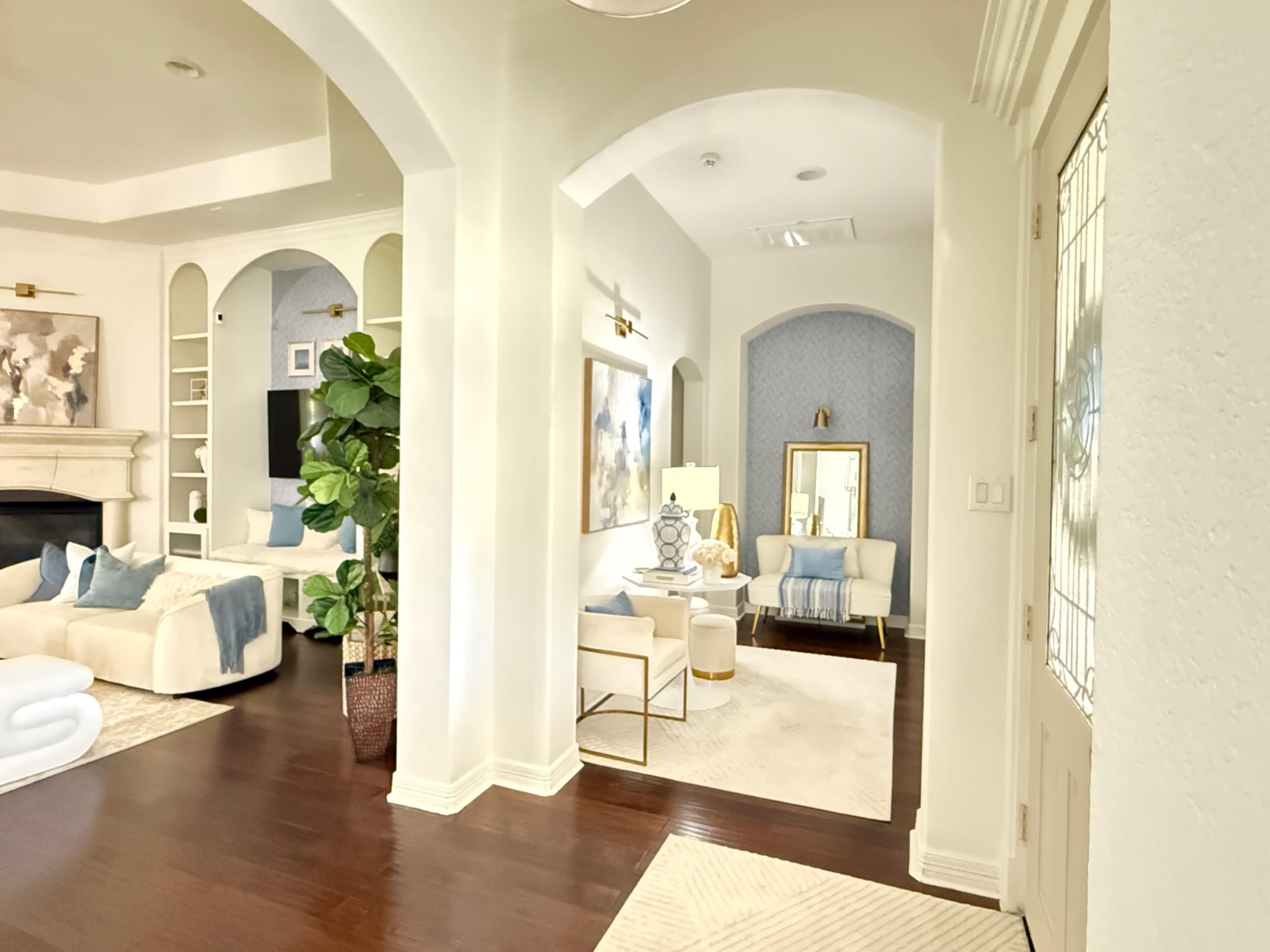 The image shows a spacious, elegantly designed living area with light-colored furniture, a decorative plant, and a contrasting dark wooden floor.