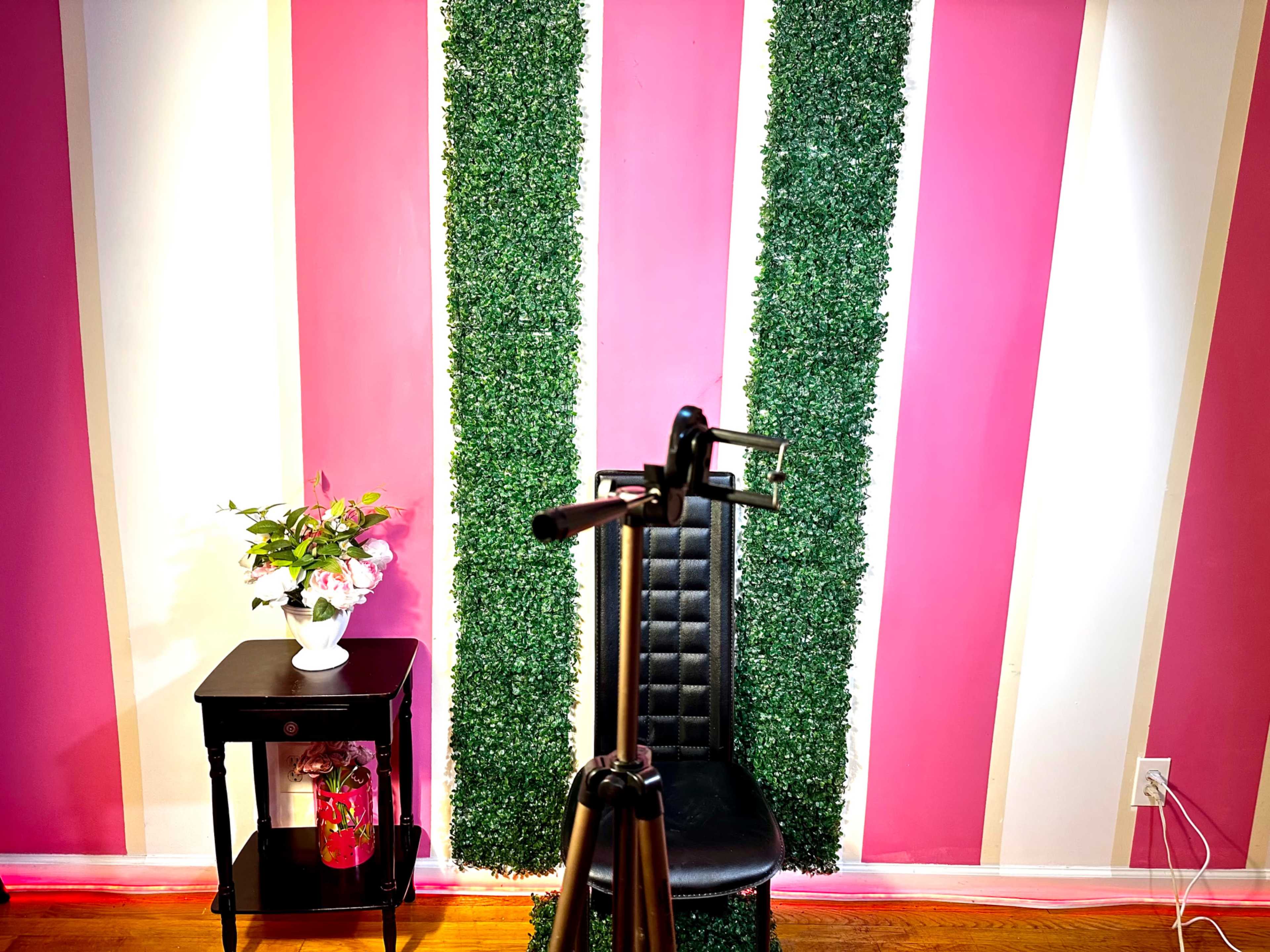 The image shows a black chair positioned in front of a pink and green striped wall, with a small table and a bouquet of flowers nearby.