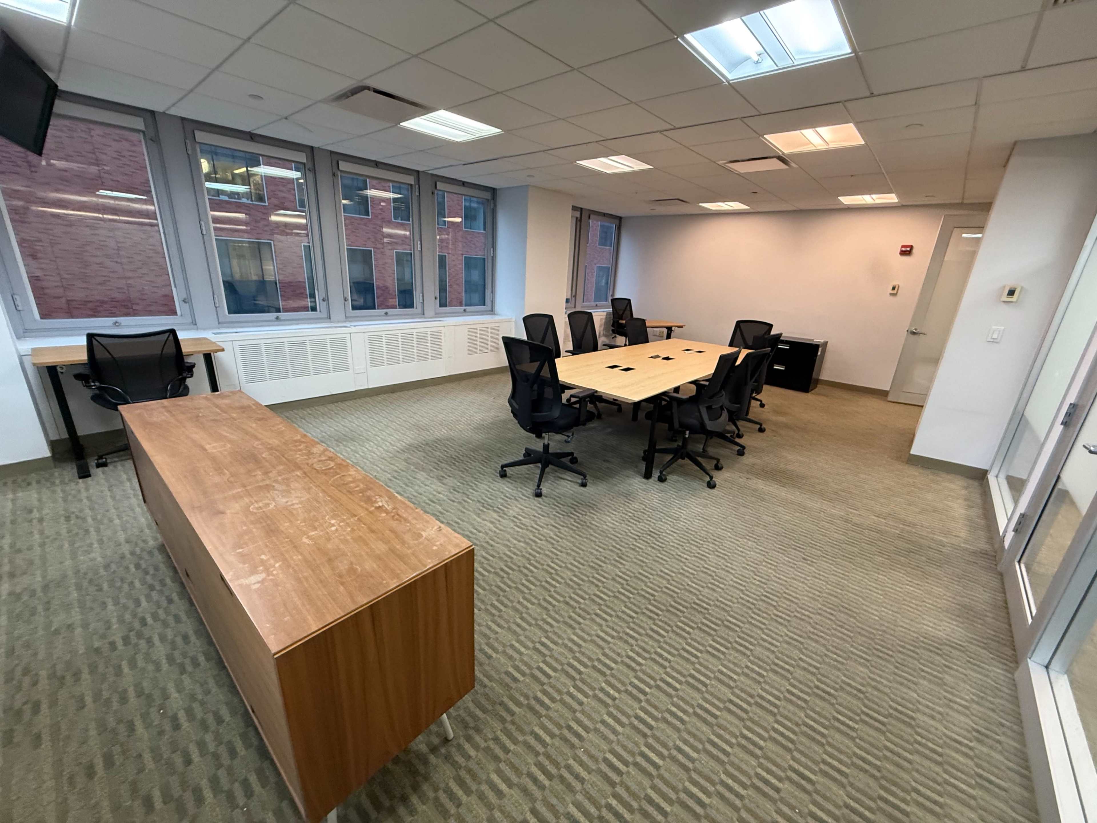 The image shows a conference room with a large wooden table surrounded by black chairs, and several windows letting in natural light.