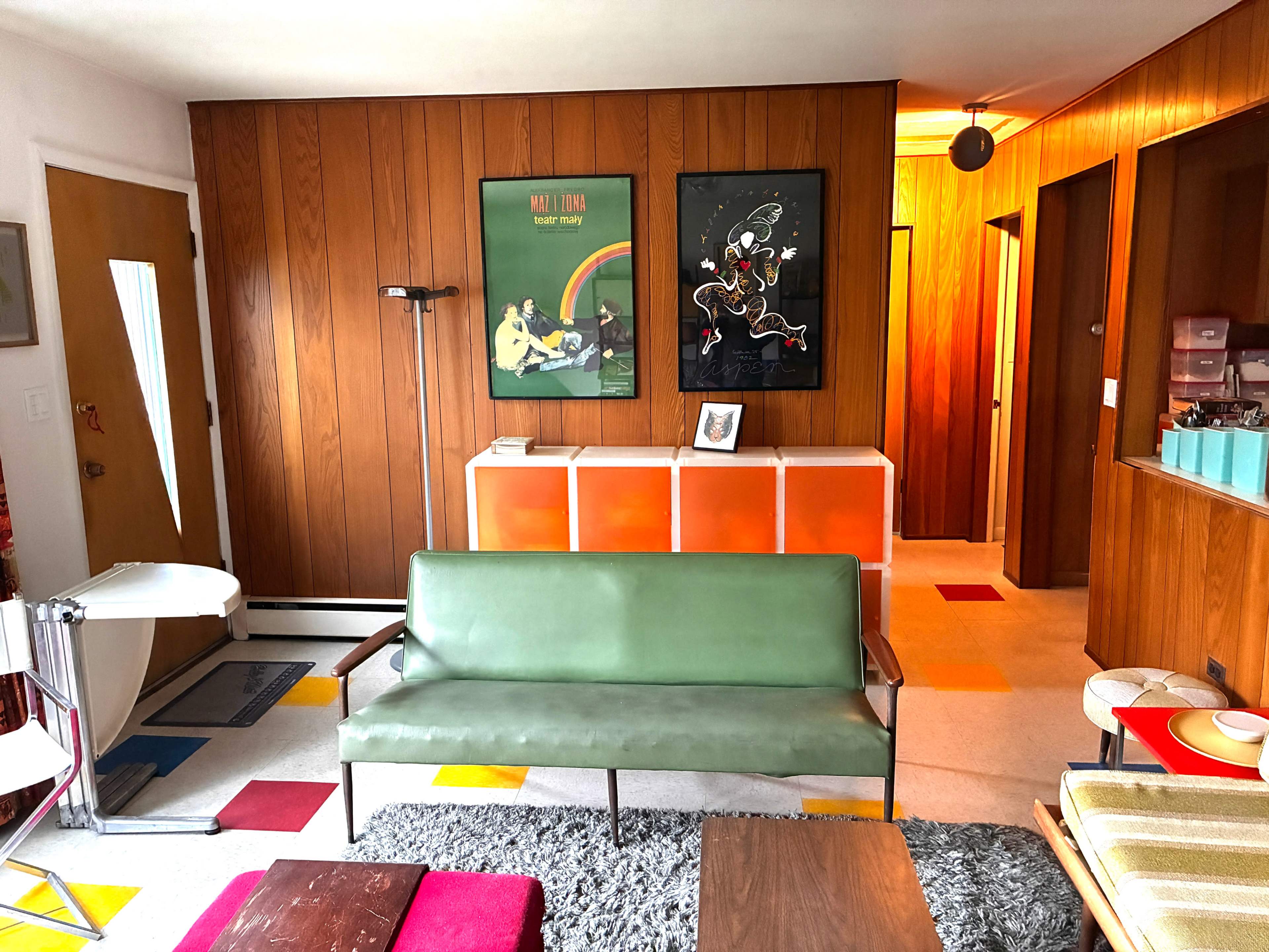 The image shows a mid-century modern living room with wooden paneling, colorful accents, and a green sofa facing a wall adorned with framed artwork.
