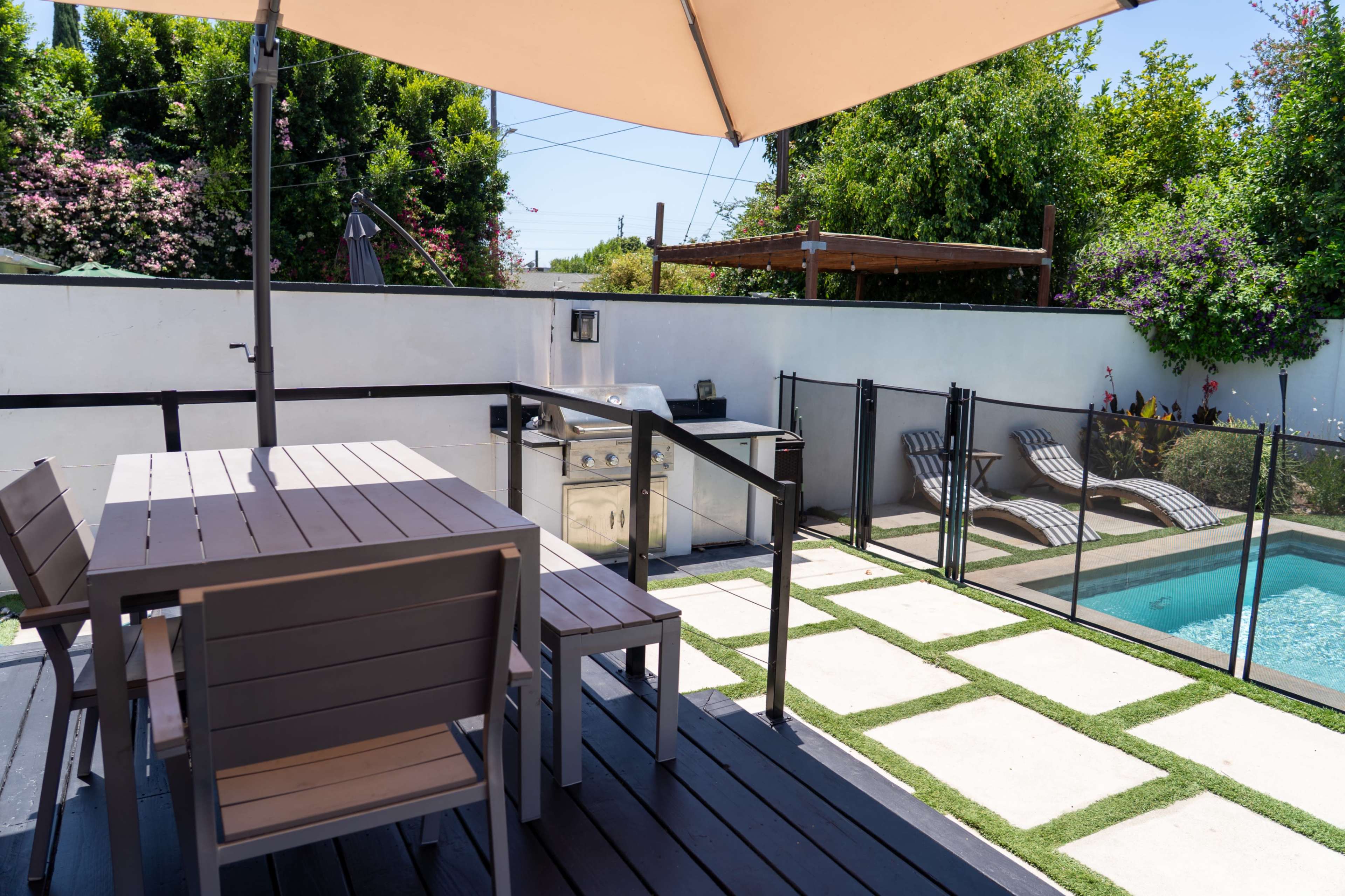 A patio area features a table with chairs, a grill, and a swimming pool surrounded by grass and paving stones.