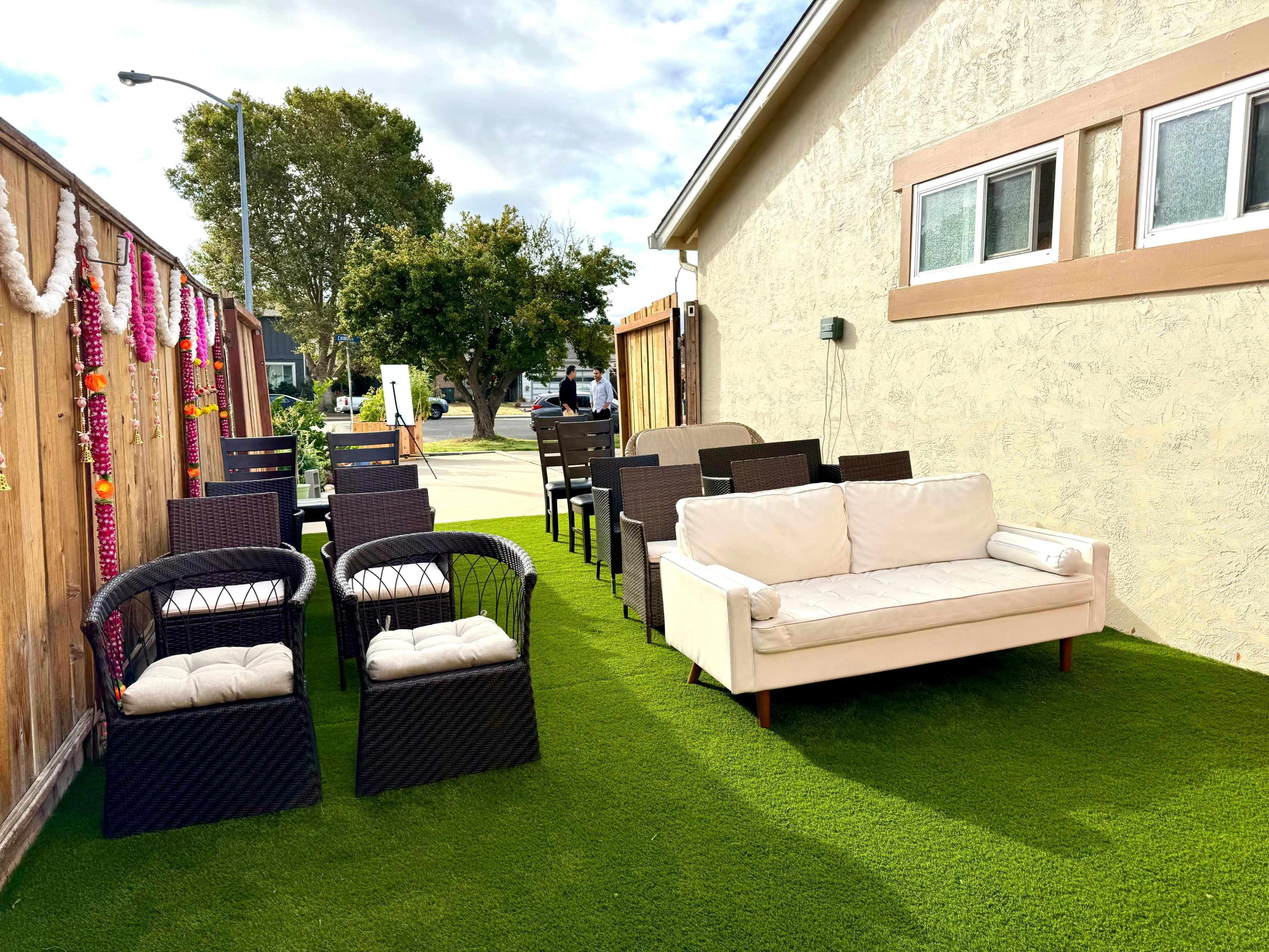 An outdoor seating area with several chairs and a white couch arranged on artificial grass next to a fence adorned with decorative garlands.
