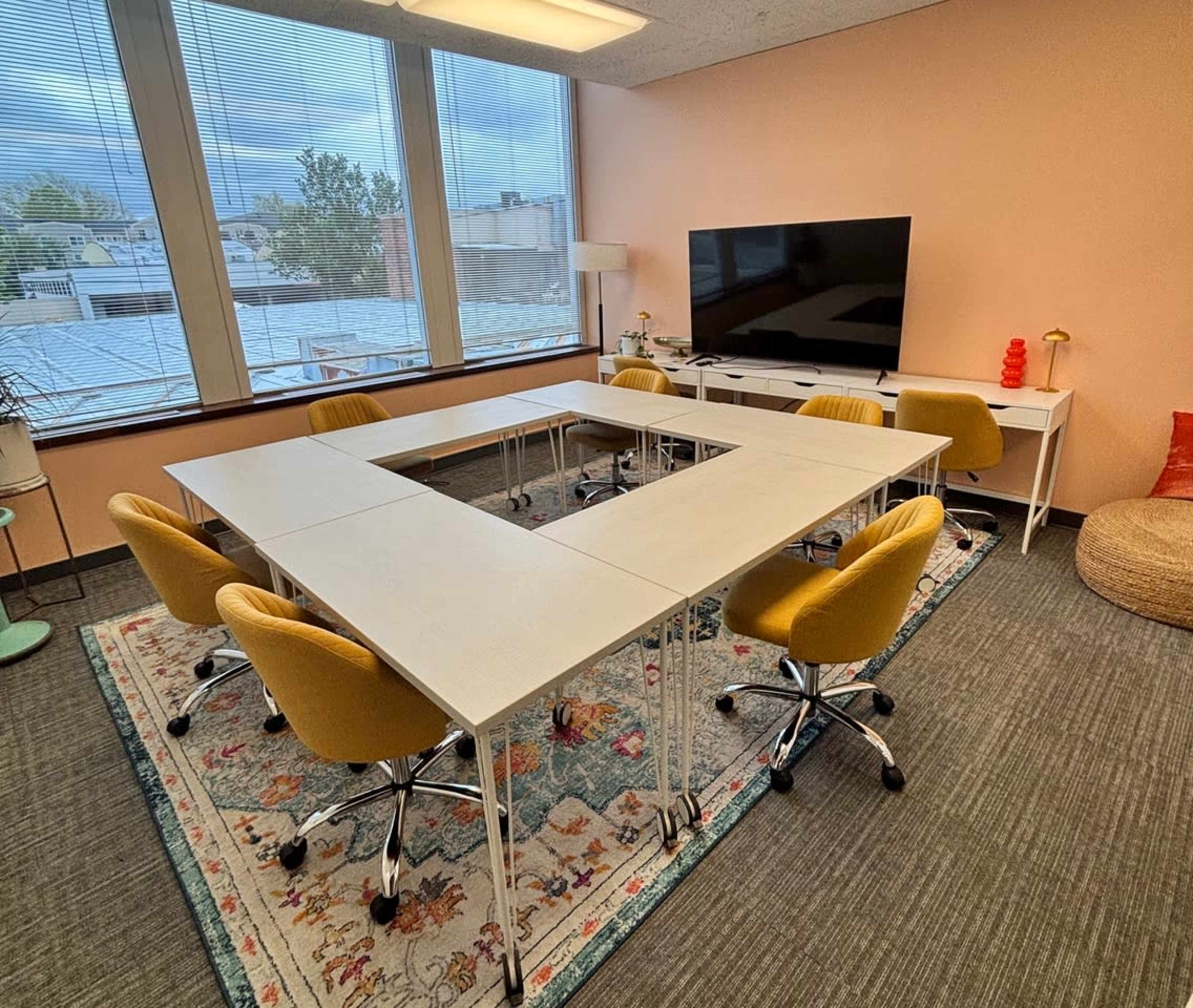 The image shows a modern meeting room with a rectangular arrangement of white tables surrounded by yellow office chairs, a large television, and a decorative rug.