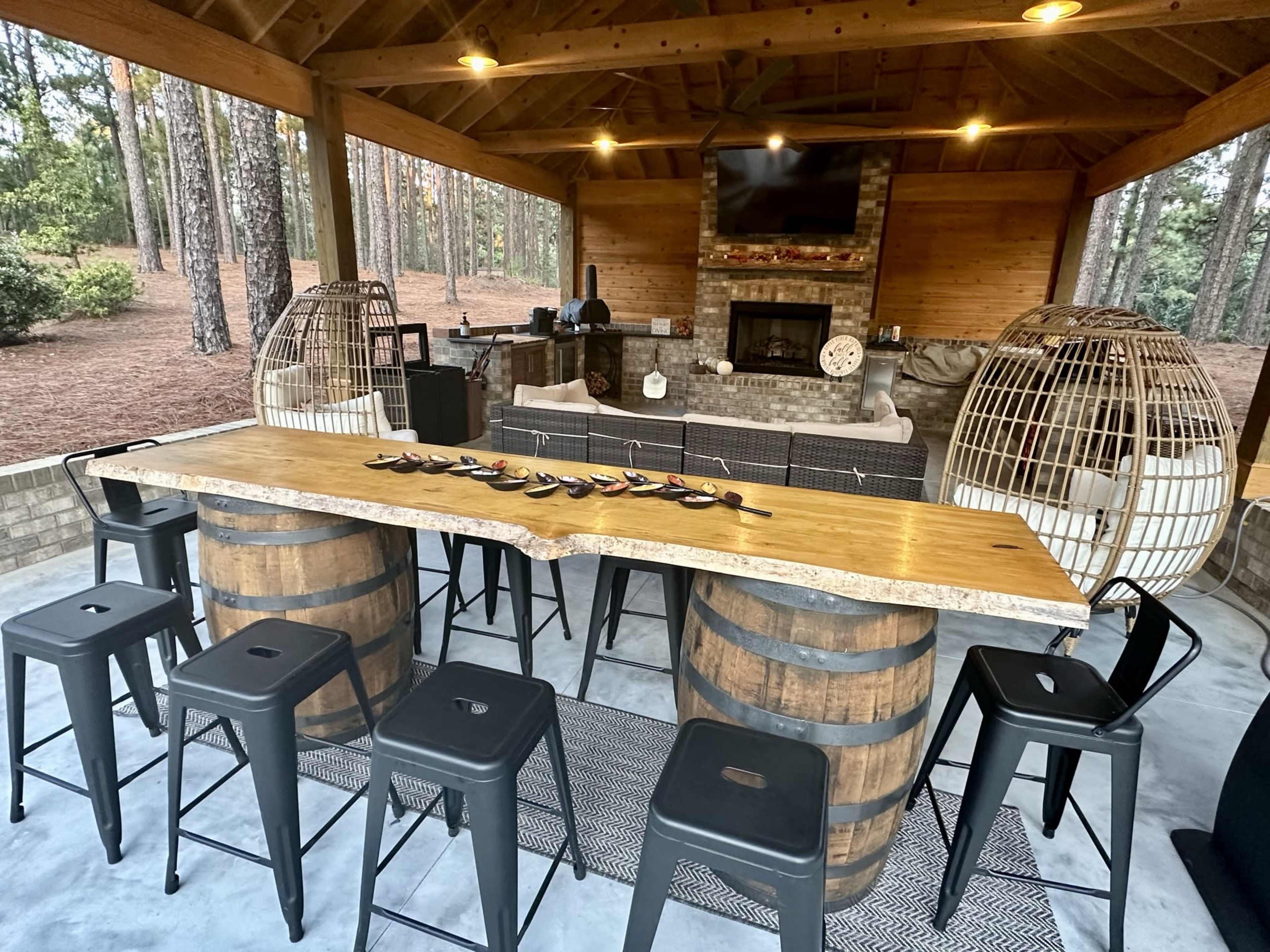 An outdoor bar area with a wooden table supported by barrel bases, surrounded by metal stools, and a stone fireplace in the background.