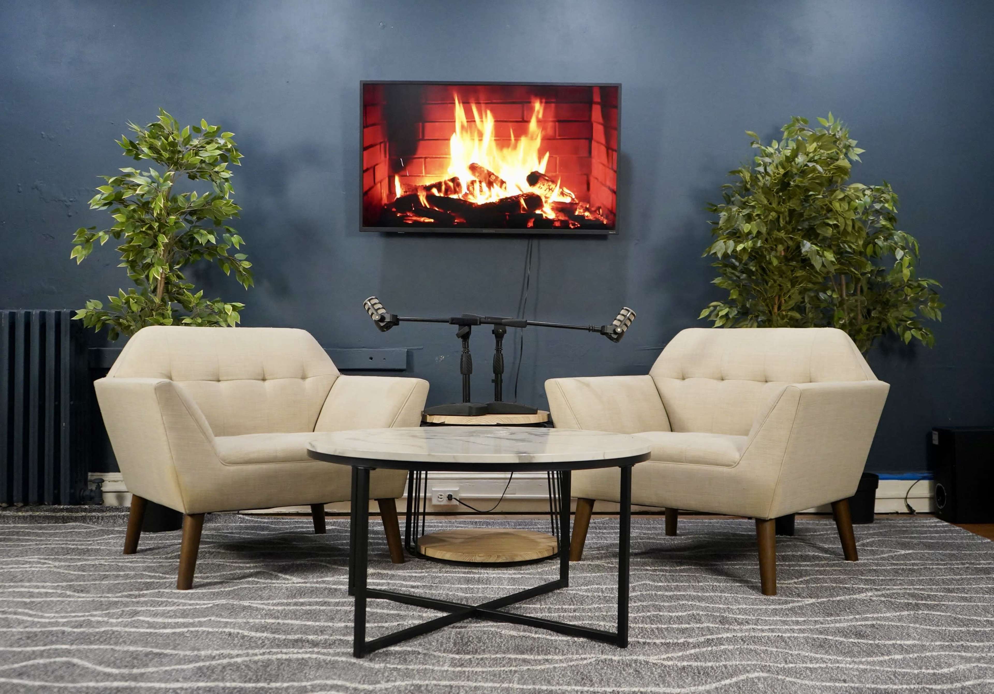 The image features two beige armchairs facing a round table, with an artificial fireplace displayed on a wall-mounted screen in a room adorned with potted plants.