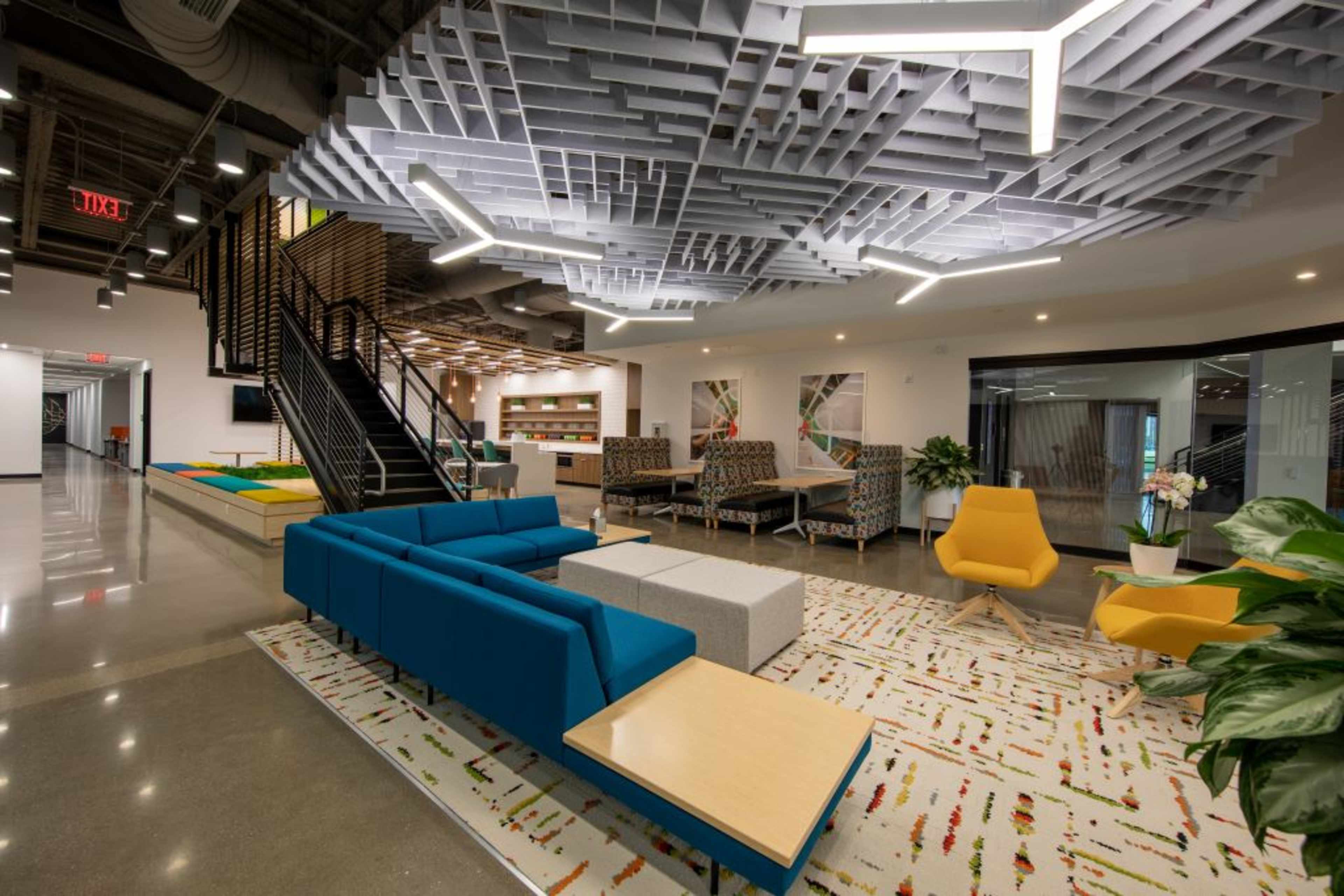 The image shows a modern open-office lounge area with colorful seating, a large rug, and decorative ceiling features.