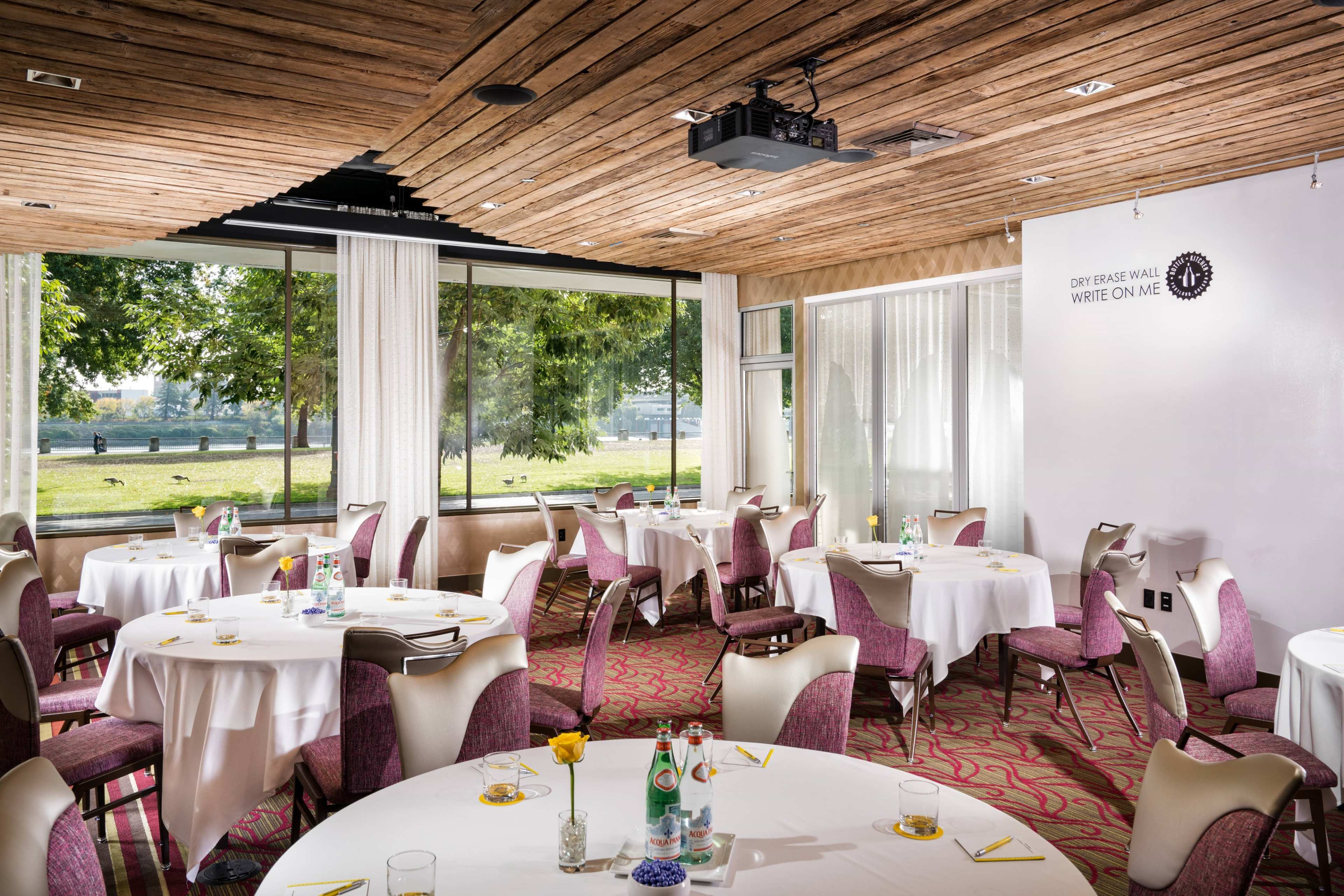 A dining area with round tables covered in white tablecloths, surrounded by chairs, and large windows overlooking a green landscape.