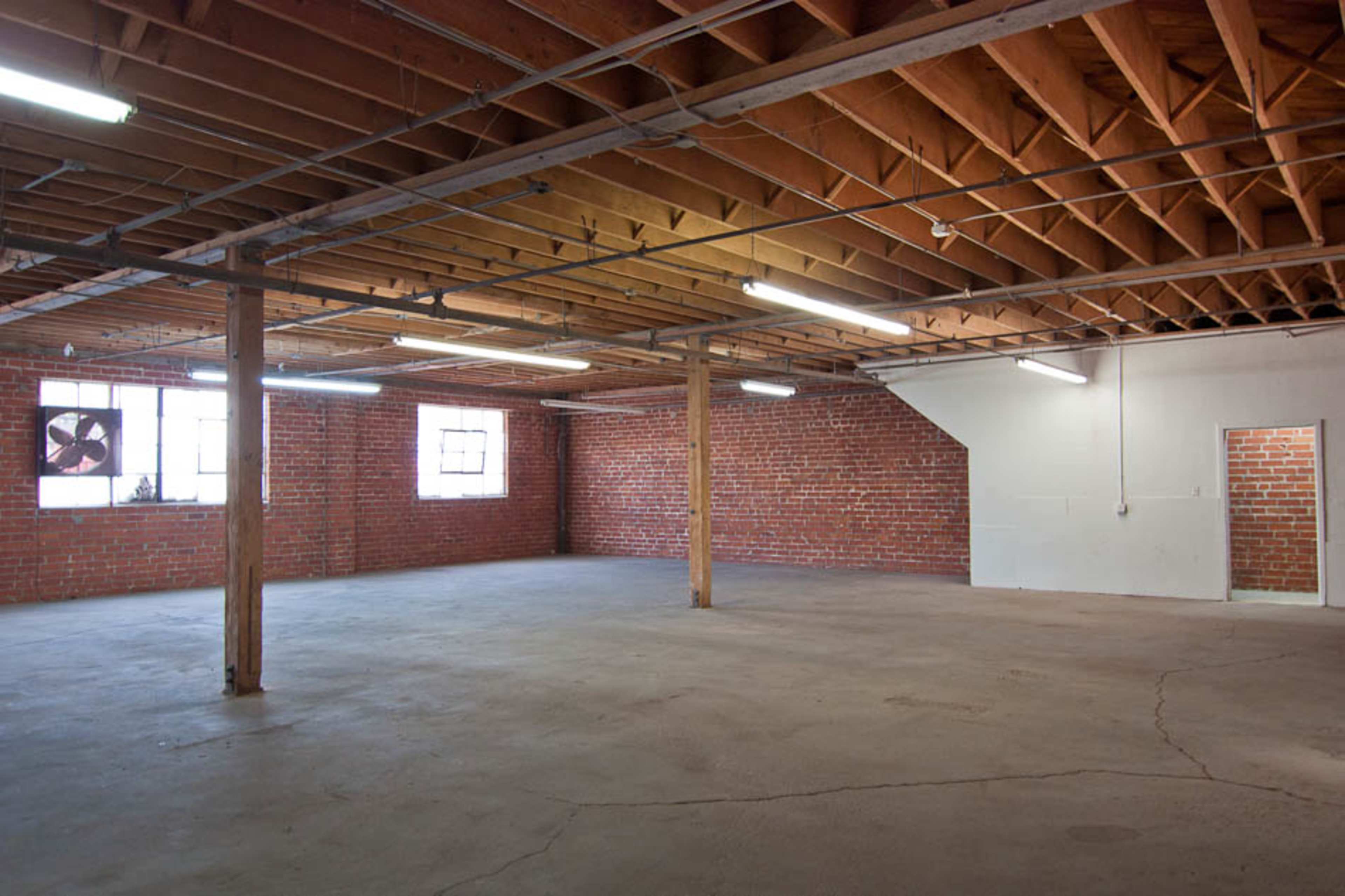 An empty industrial space with exposed wooden beams, brick walls, and concrete flooring, featuring several windows and a small door on one side.