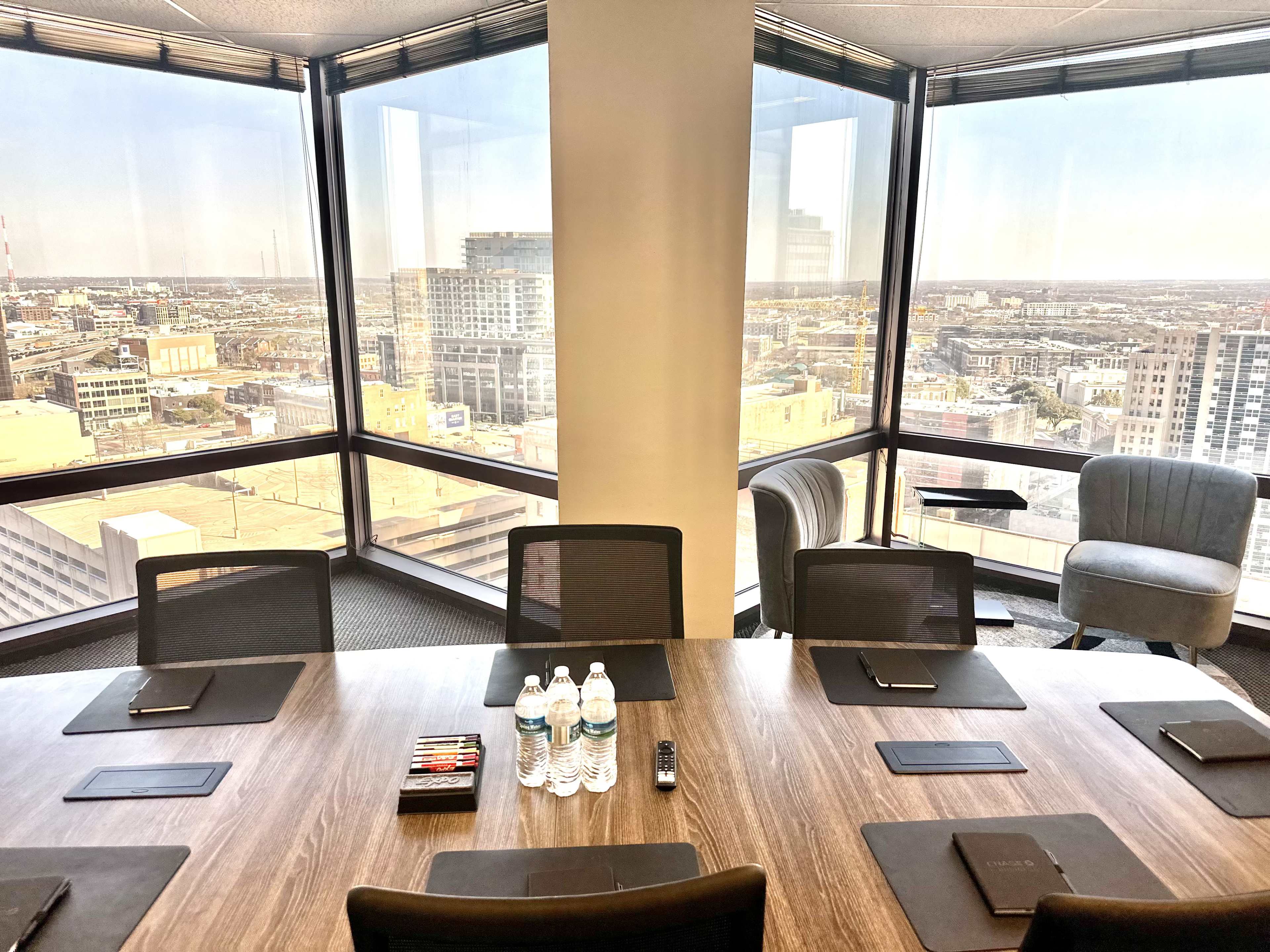 A conference room features a round wooden table with several water bottles and writing materials, overlooking a cityscape through large windows.