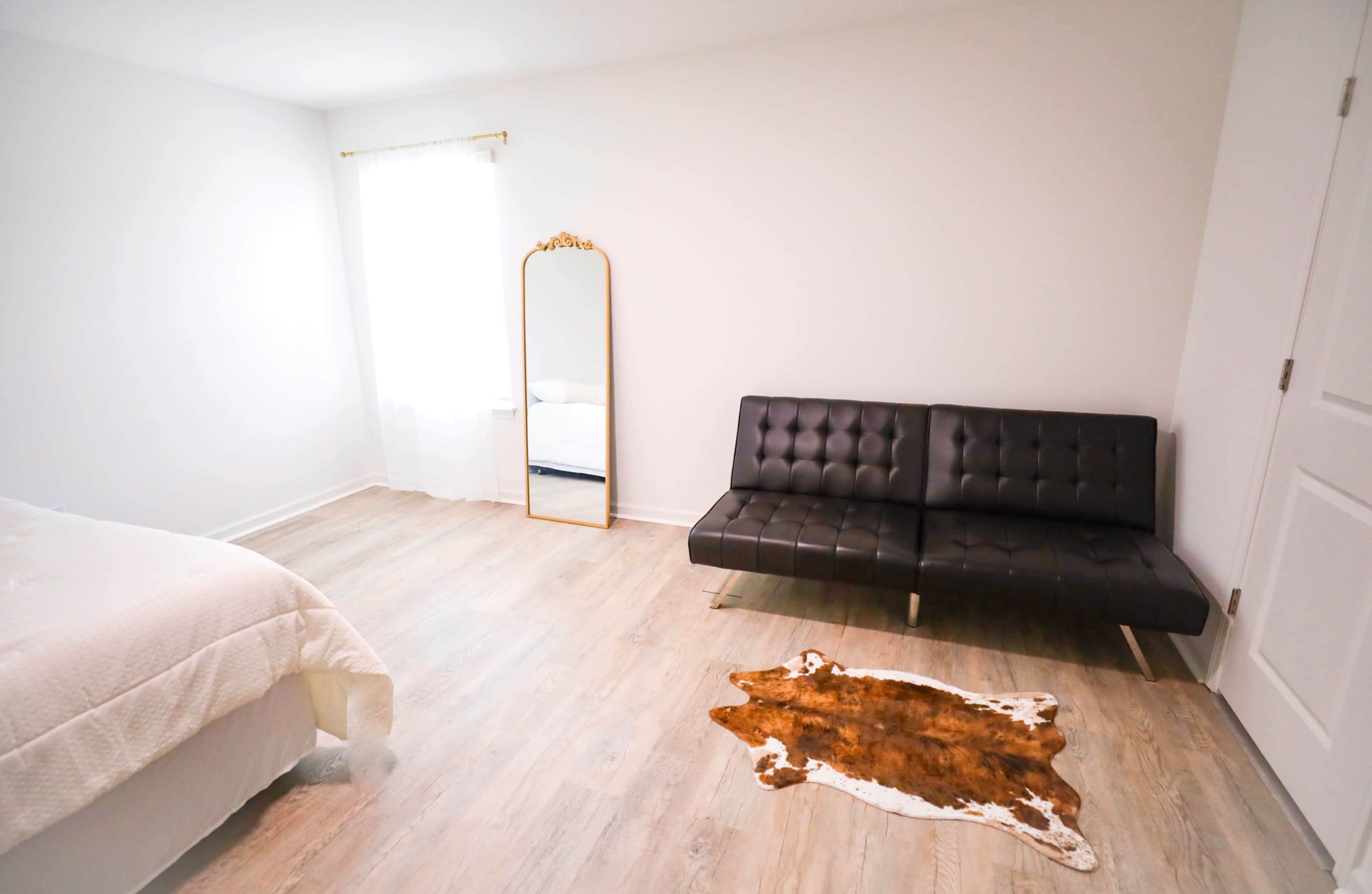 The image shows a minimalistic bedroom featuring a bed, a black sofa, a large mirror, and a cowhide rug on the floor.
