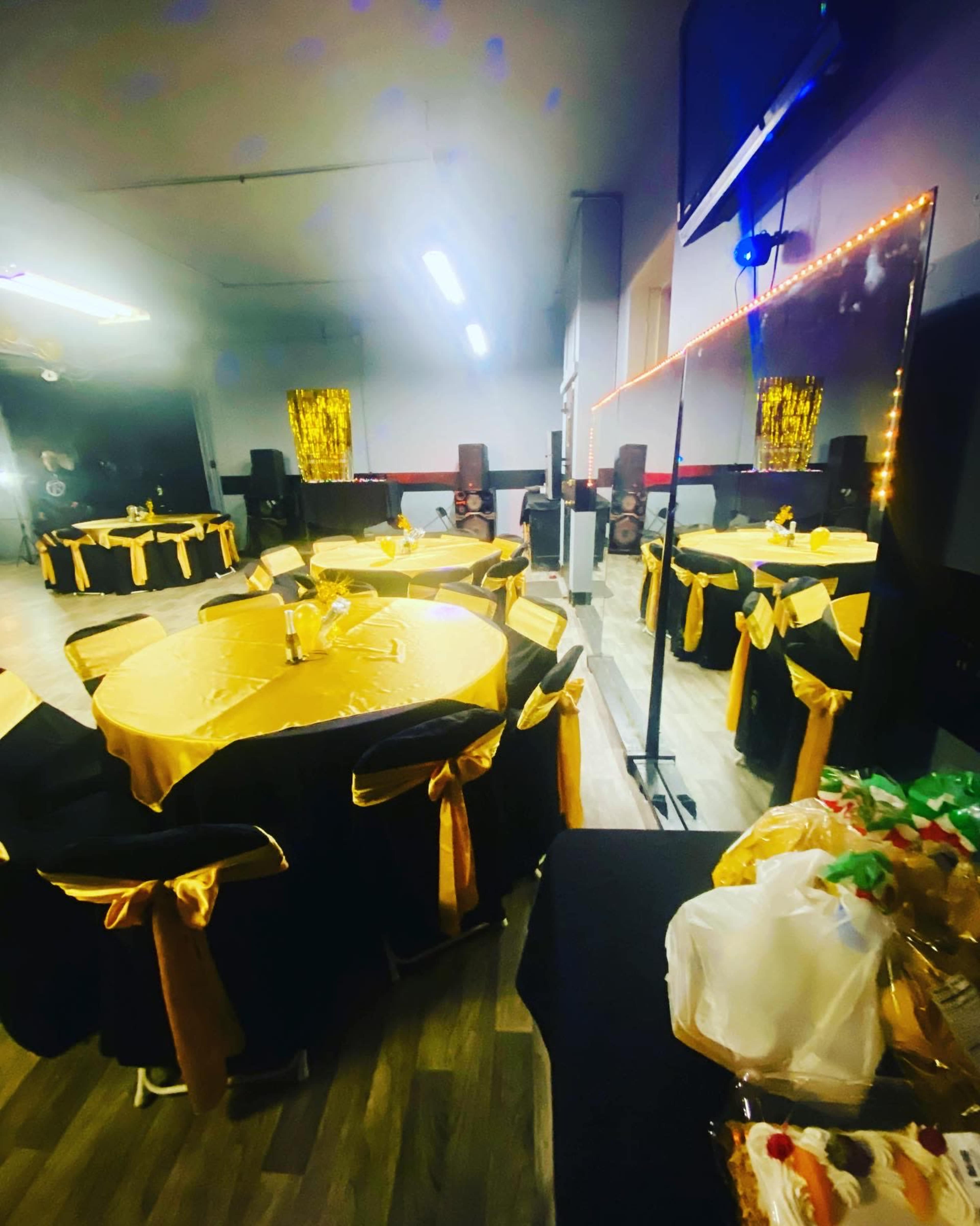 The scene shows a decorated event space featuring several round tables covered with golden tablecloths and black chairs adorned with gold sashes.
