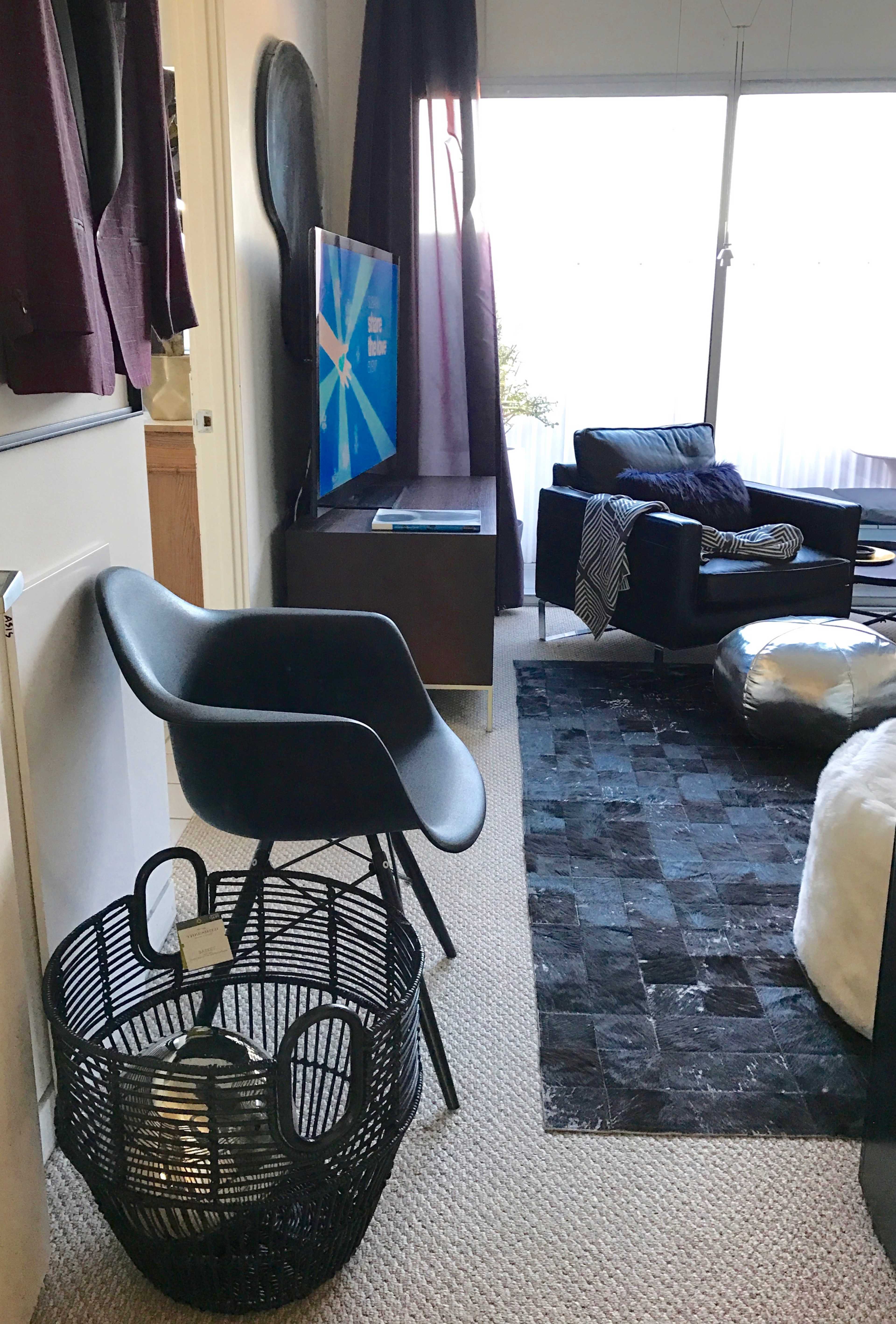The image shows a stylish living space featuring a black chair on a textured rug, a television on a cabinet, and a dark sofa beside a window with sheer curtains.