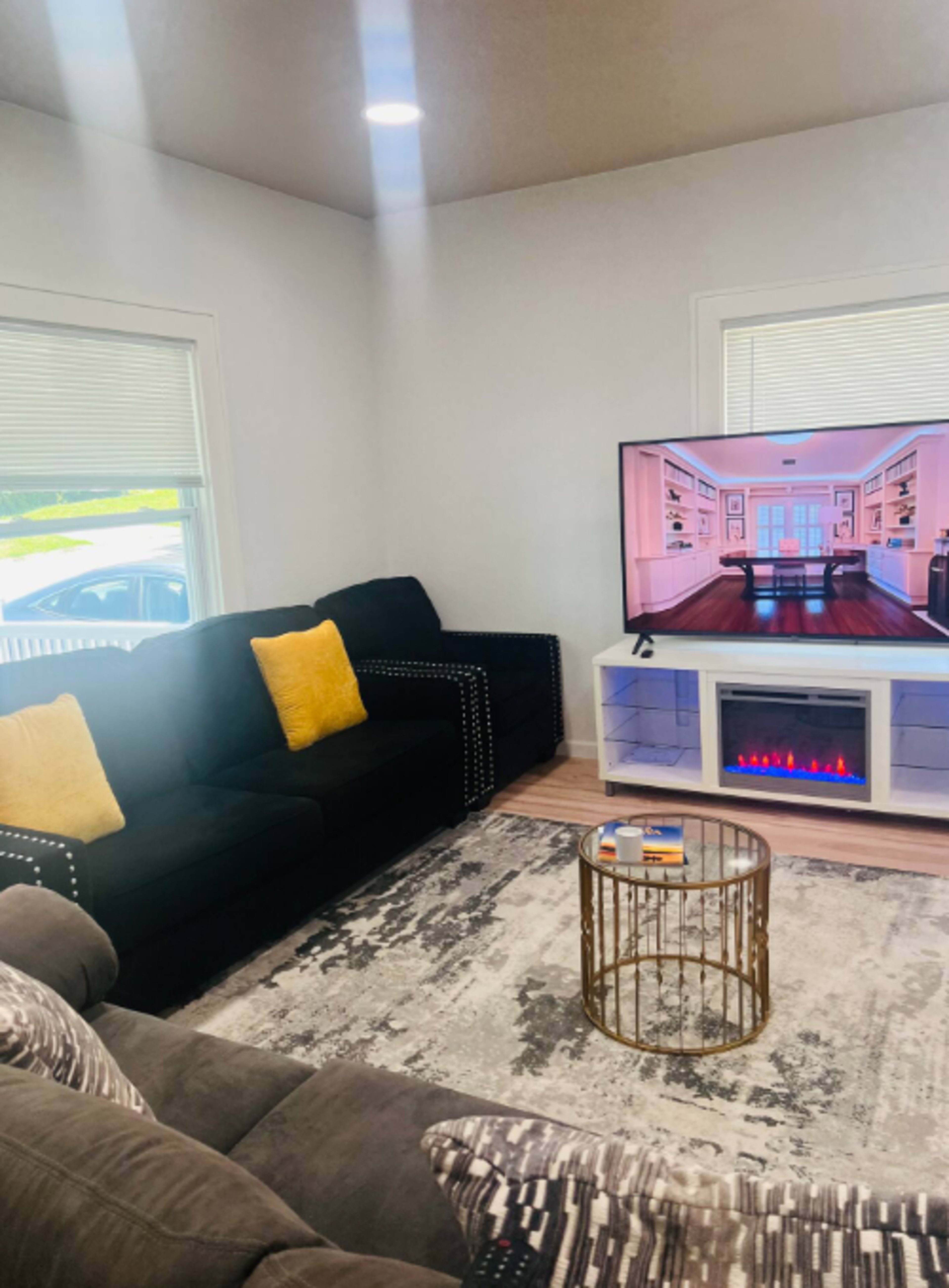 The living room features a dark sofa with yellow cushions, a coffee table, and a large television displaying a room setting, with a faux fireplace below.