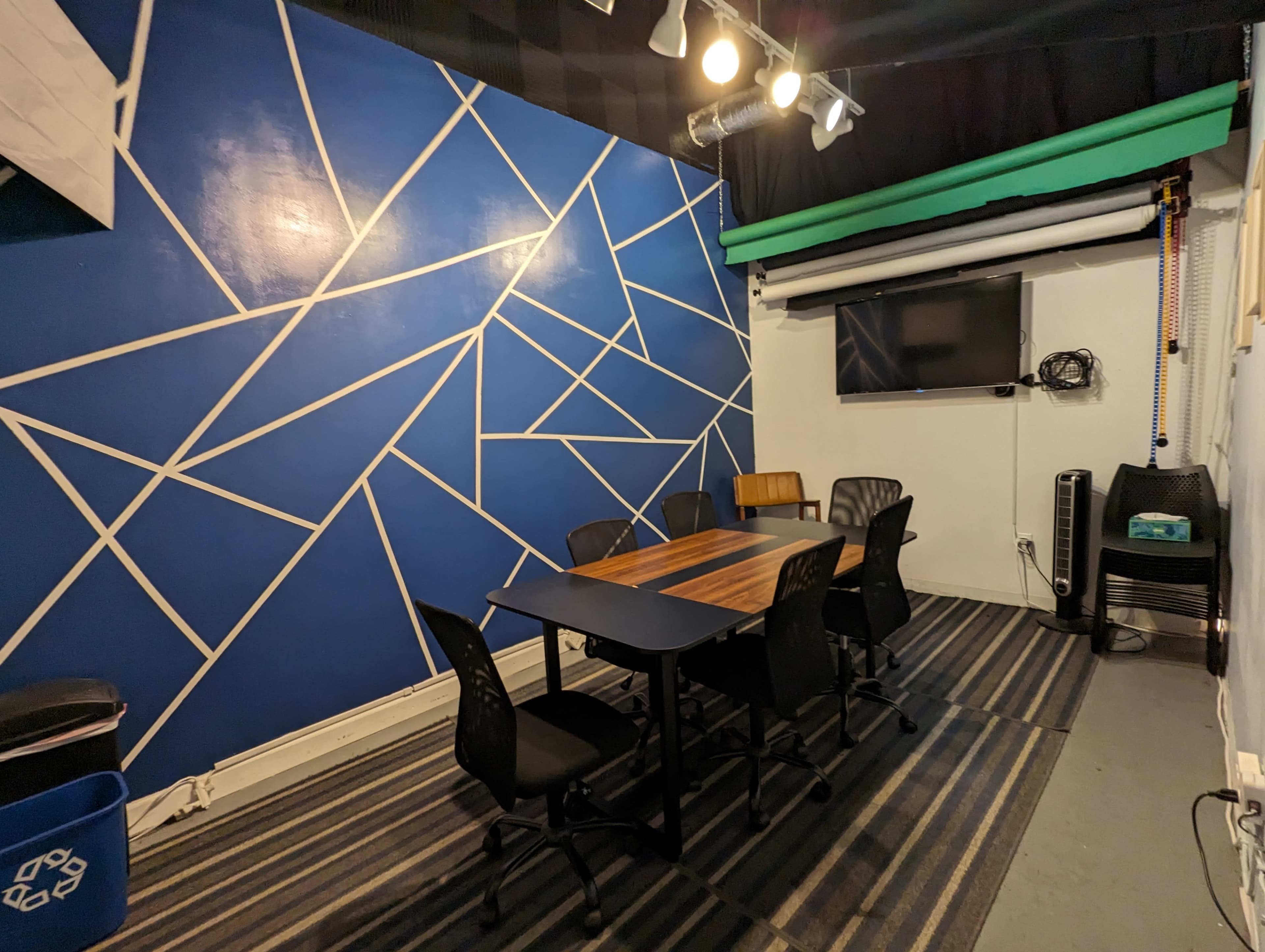 A small meeting room with a wooden table surrounded by black chairs, a blue geometric feature wall, and a television mounted on the wall.