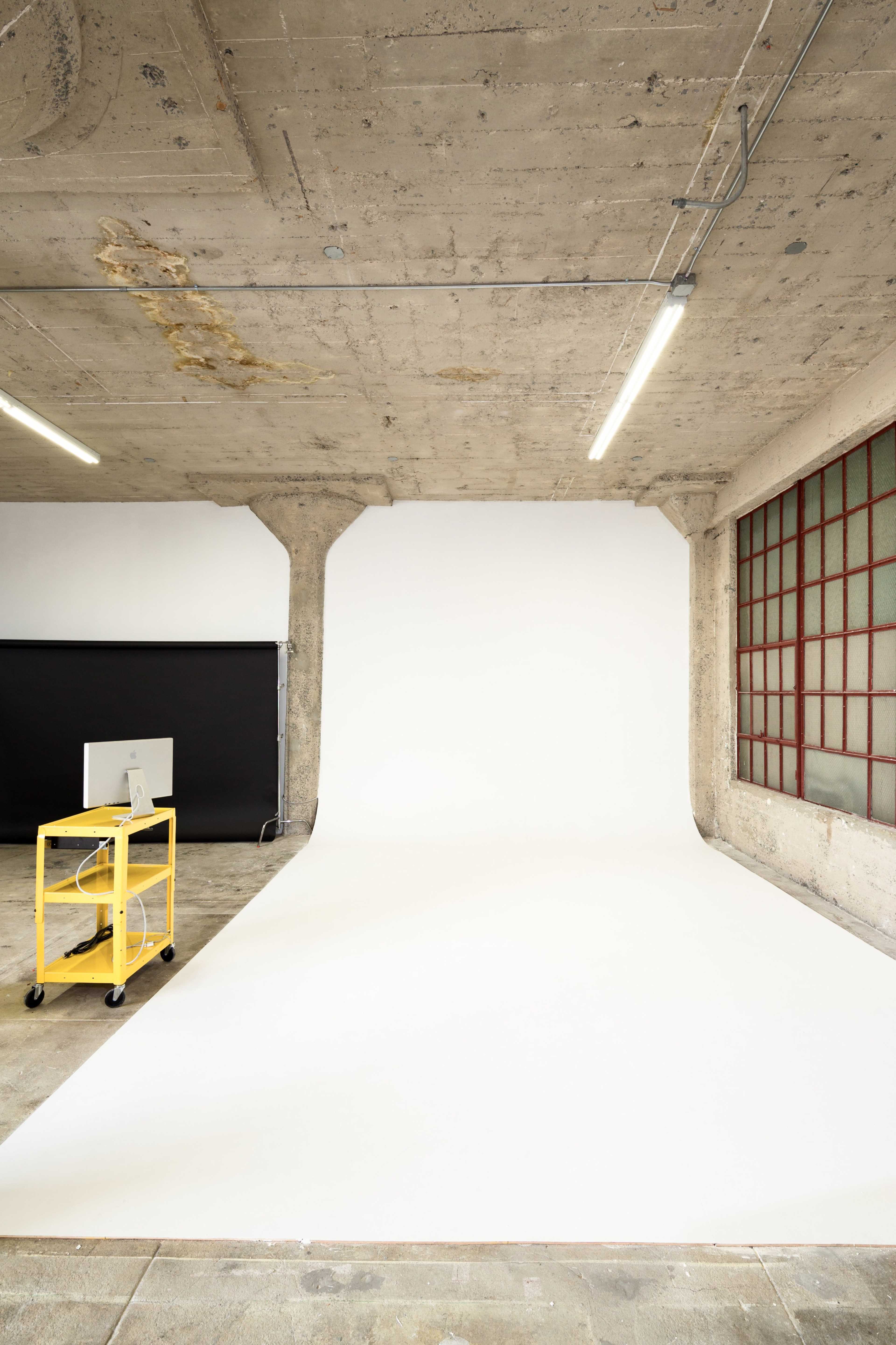 A minimalist studio space with a white backdrop, a yellow cart with a computer, and concrete walls.