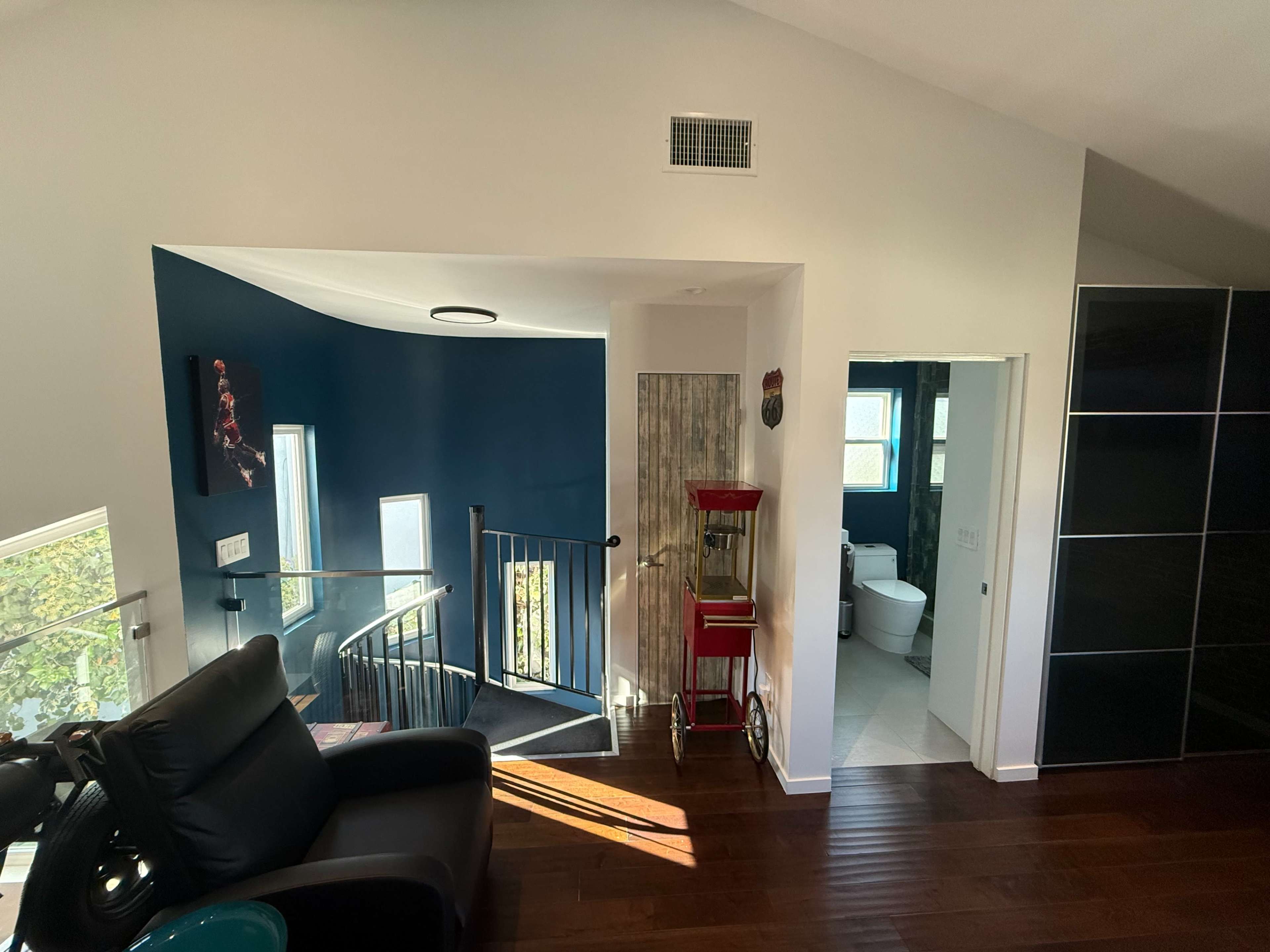 The image shows a modern interior space featuring a staircase with a railing, a dark blue wall, and a nearby bathroom door, illuminated by natural light.