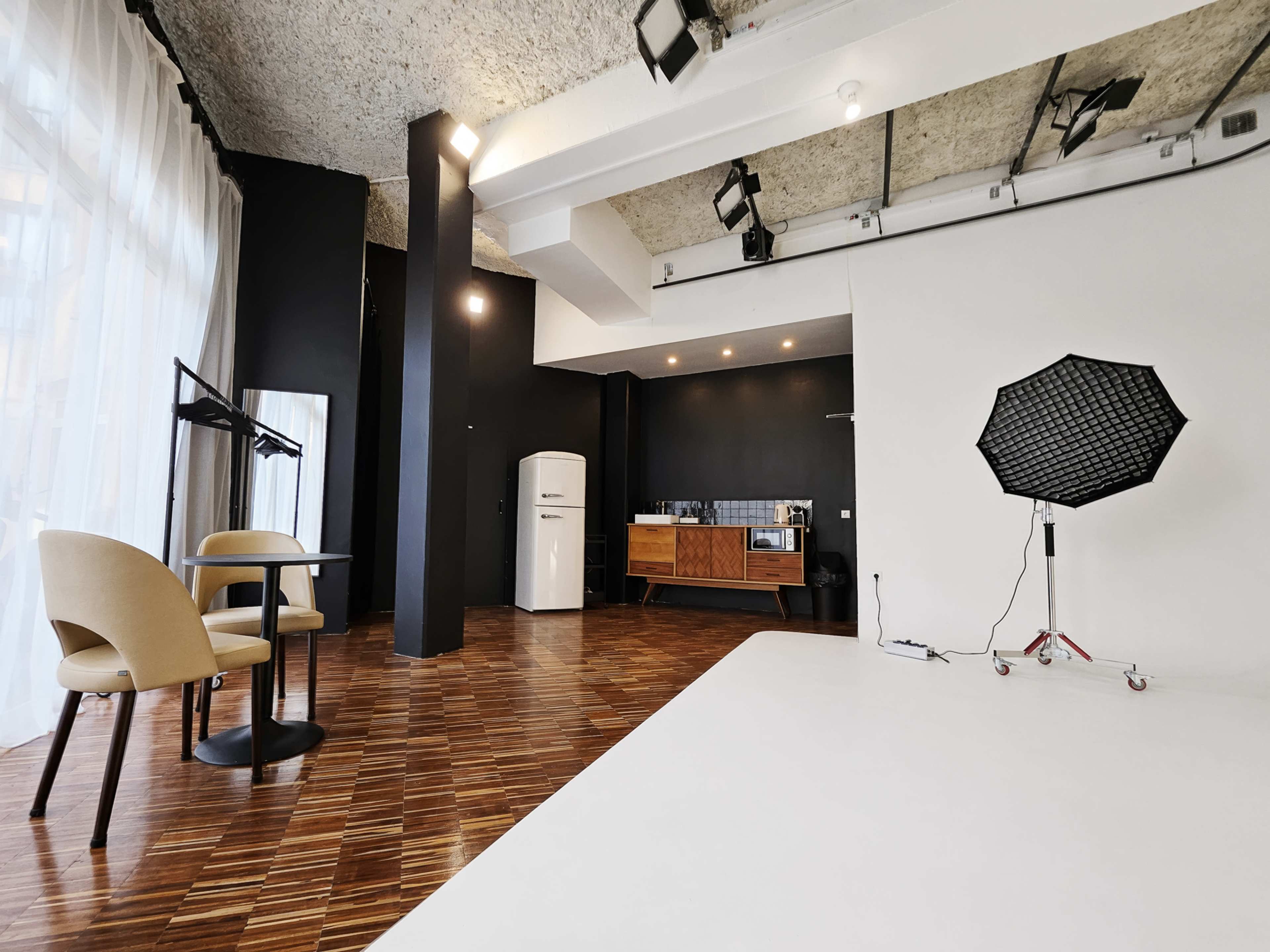 A modern photography studio features a white backdrop, a kitchenette with a refrigerator, and a table set with two chairs.