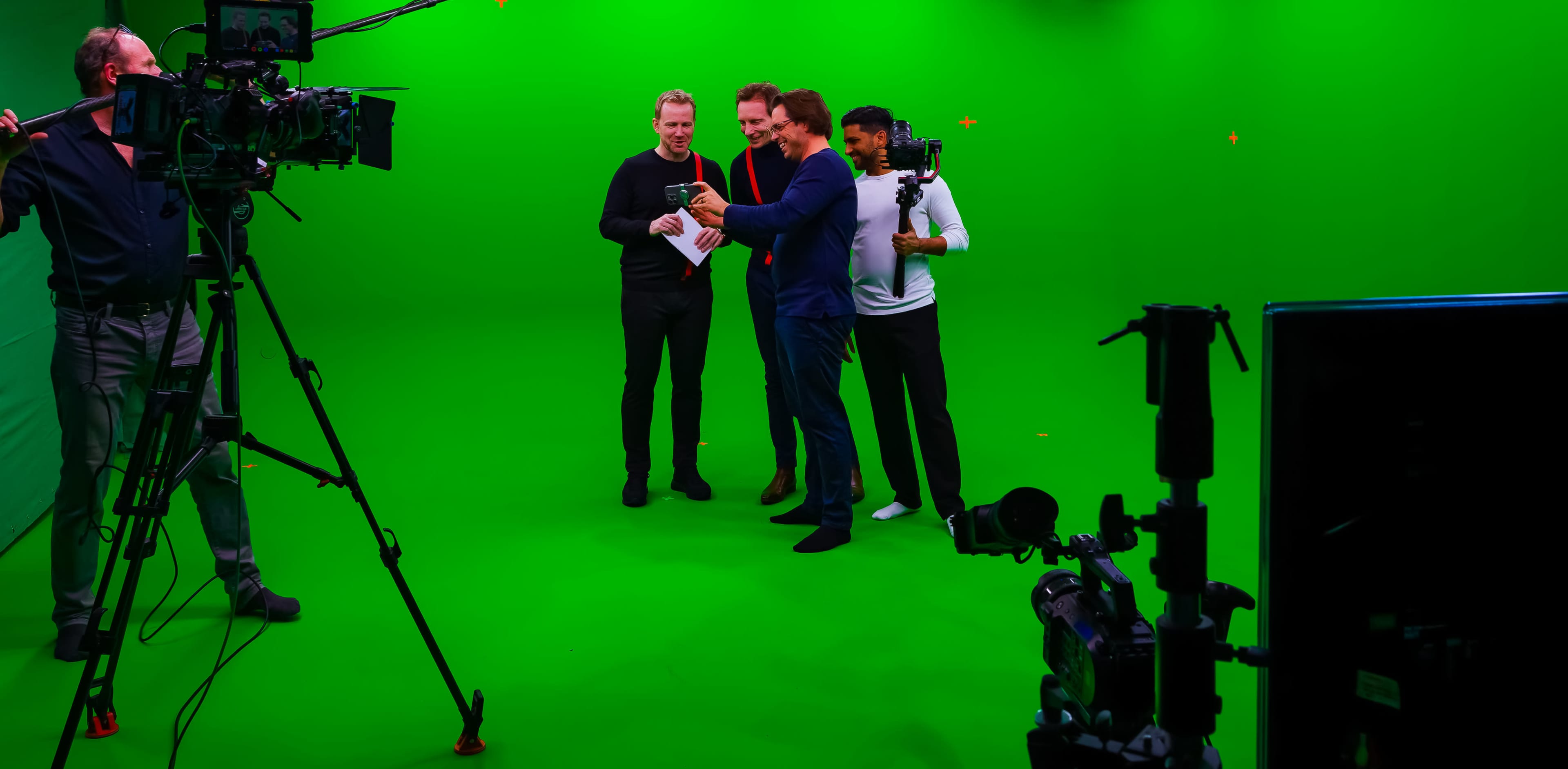 A group of four men stands in front of a green screen, with cameras and lighting equipment positioned around them.