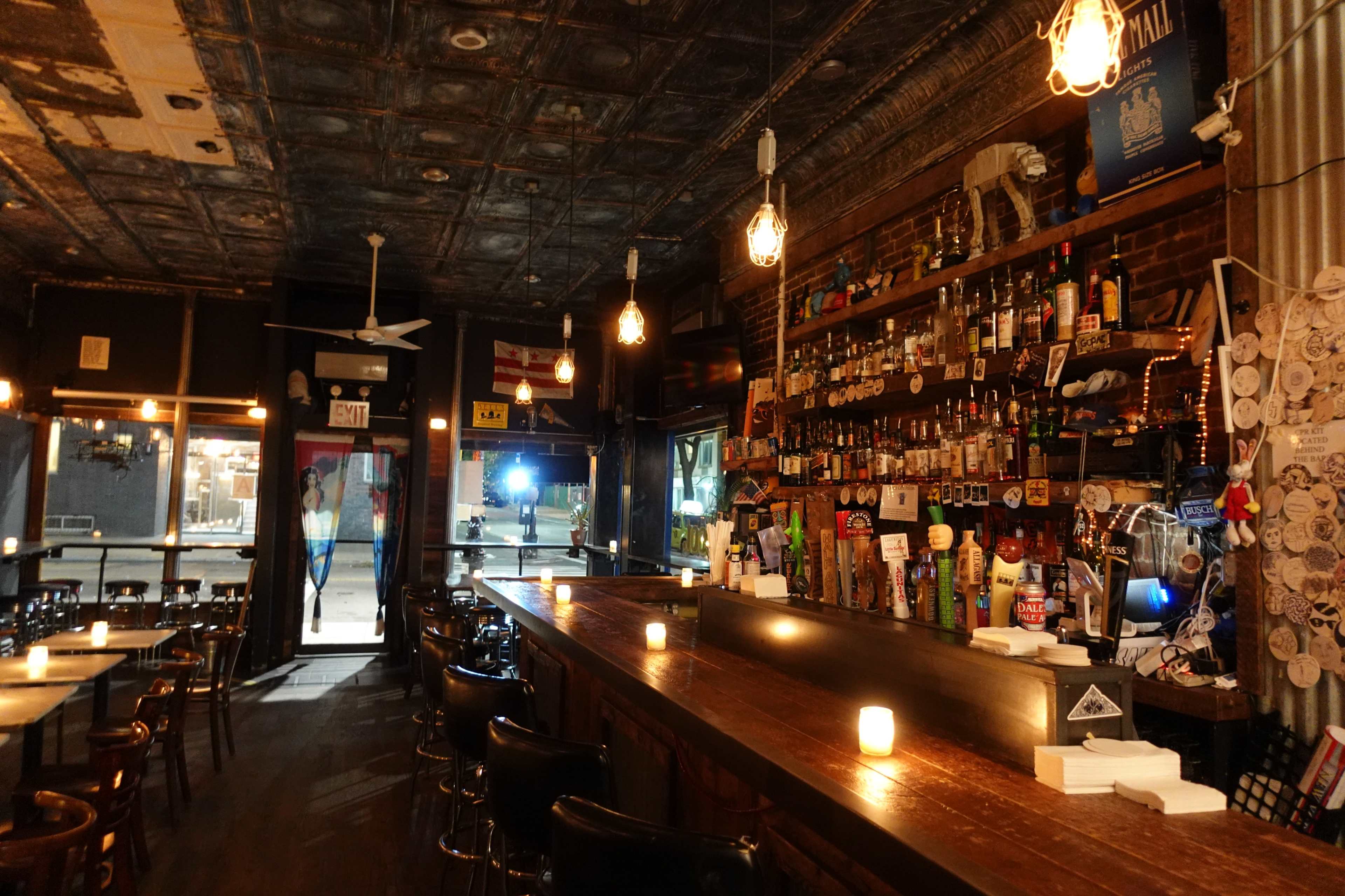 An empty bar with a long wooden counter, shelves stocked with various liquor bottles, and exposed light bulbs hanging from the ceiling.