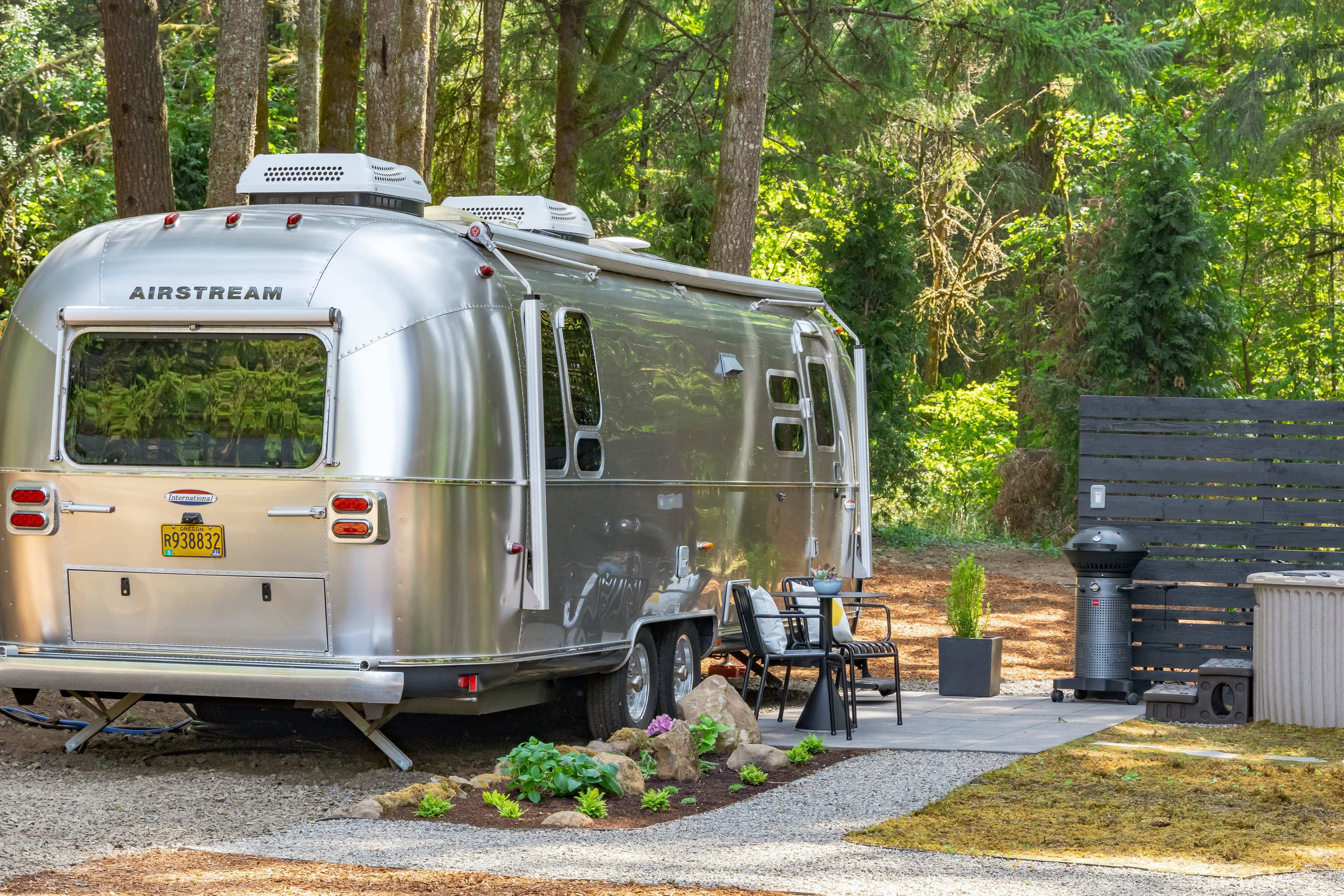 A silver Airstream trailer is parked beside a stone patio with outdoor furniture in a wooded area.