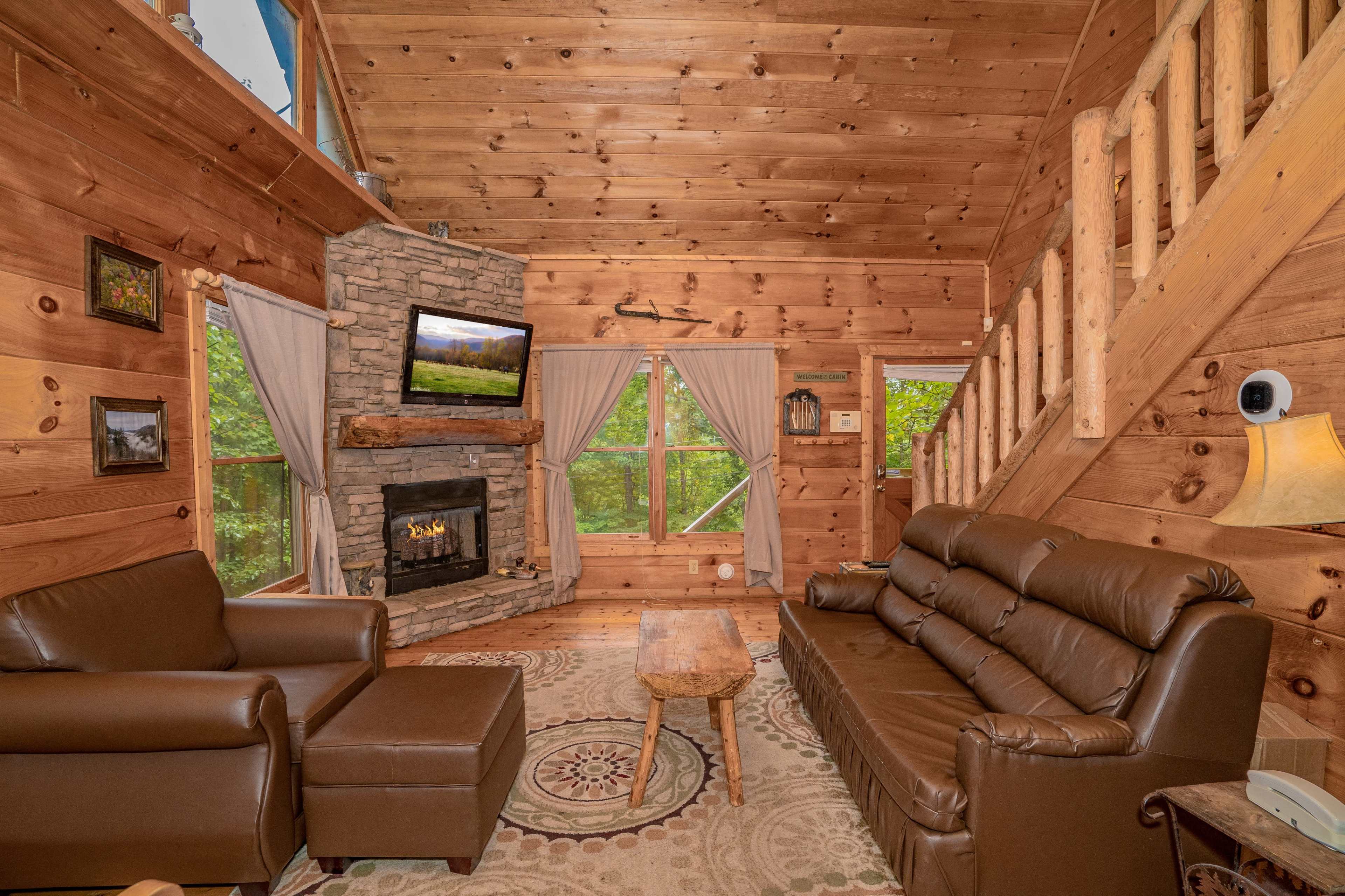 The image shows a rustic wooden living room with leather sofas, a stone fireplace, and a staircase leading to an upper level.
