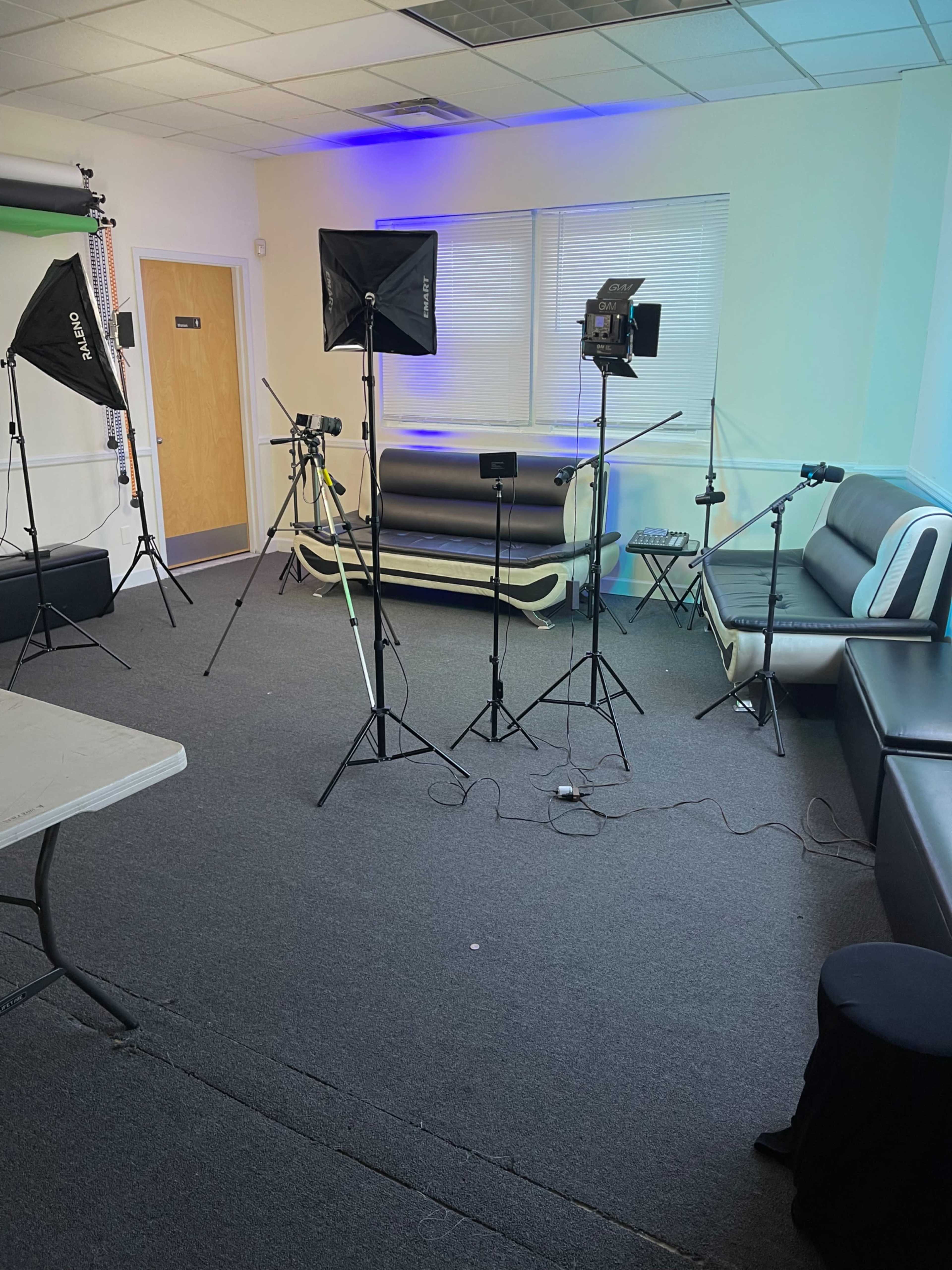 The image shows a studio setup with multiple lighting equipment, a table, and a black and white couch in a room with light blue accents.