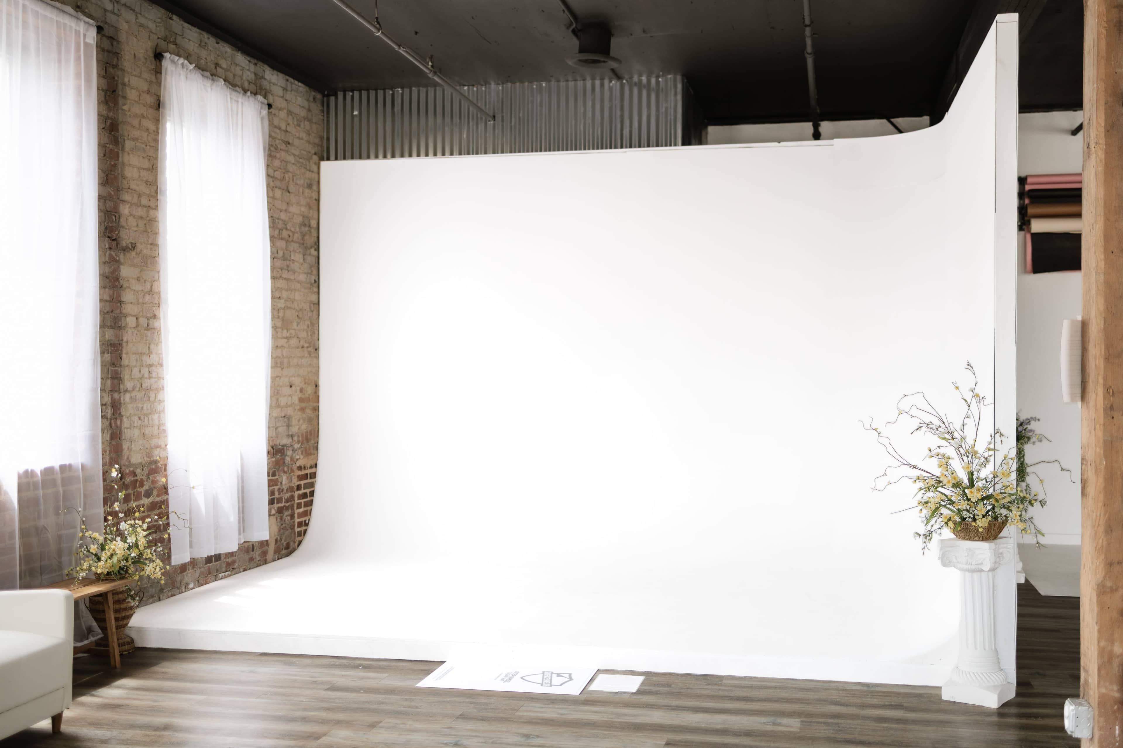 The image shows a bright studio space with a white backdrop, wooden floors, and large windows allowing natural light to enter.