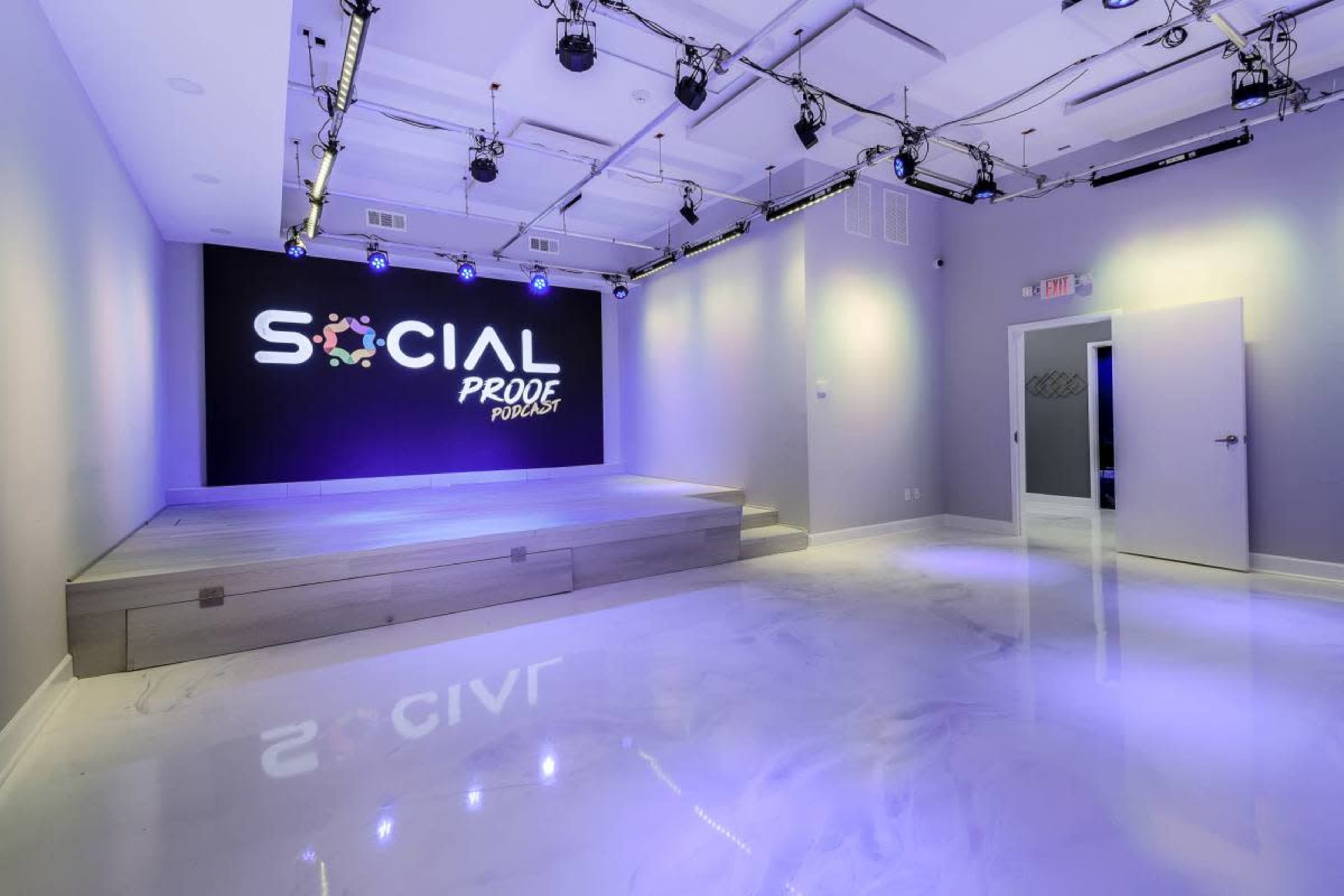 The image shows a modern event space with a raised stage featuring a lit backdrop that says "SOCIAL PROOF," and polished concrete floors under bright lighting.