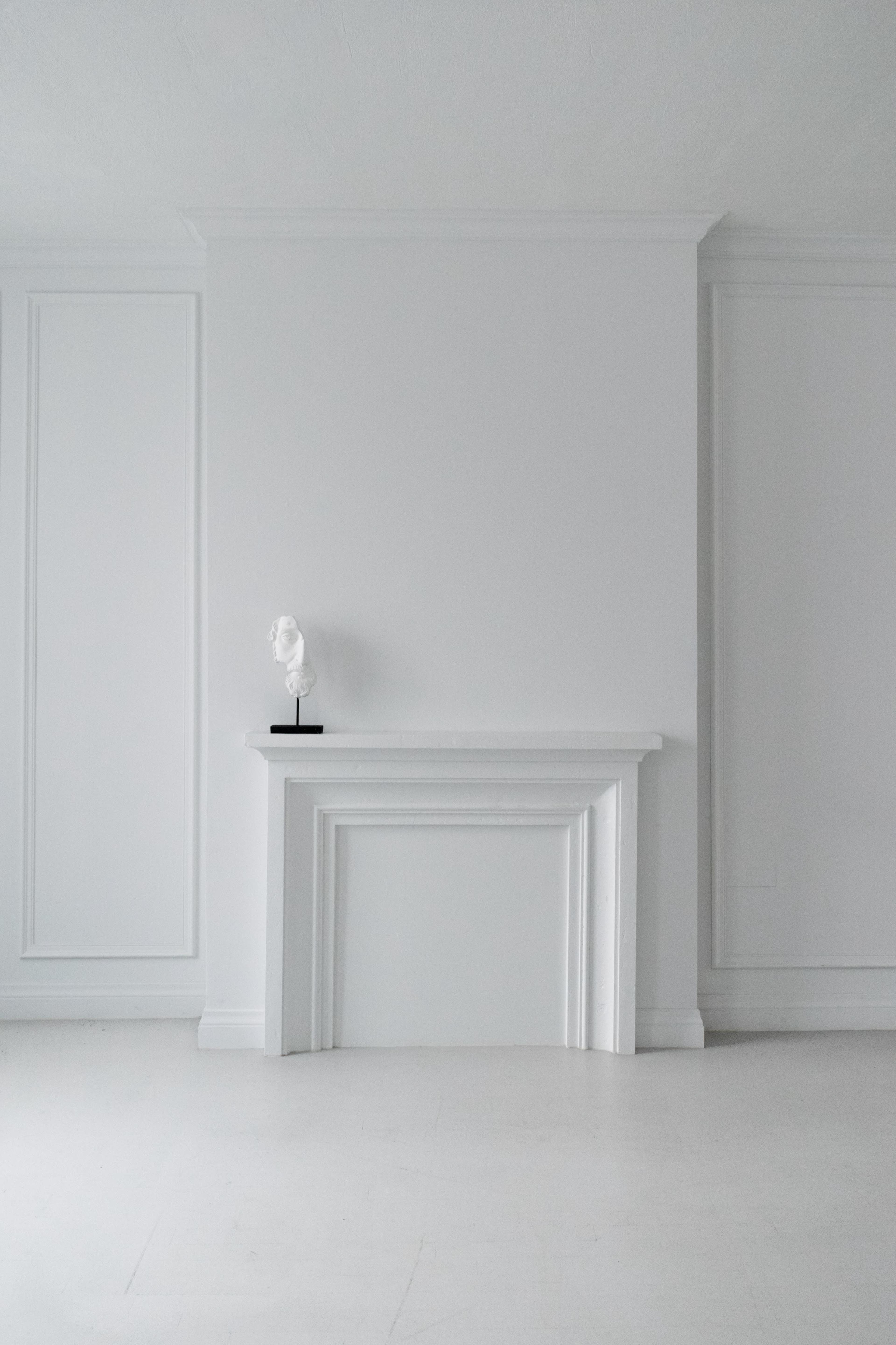 A minimalist room with white walls, featuring a simple white mantel and a sculpture on a stand.