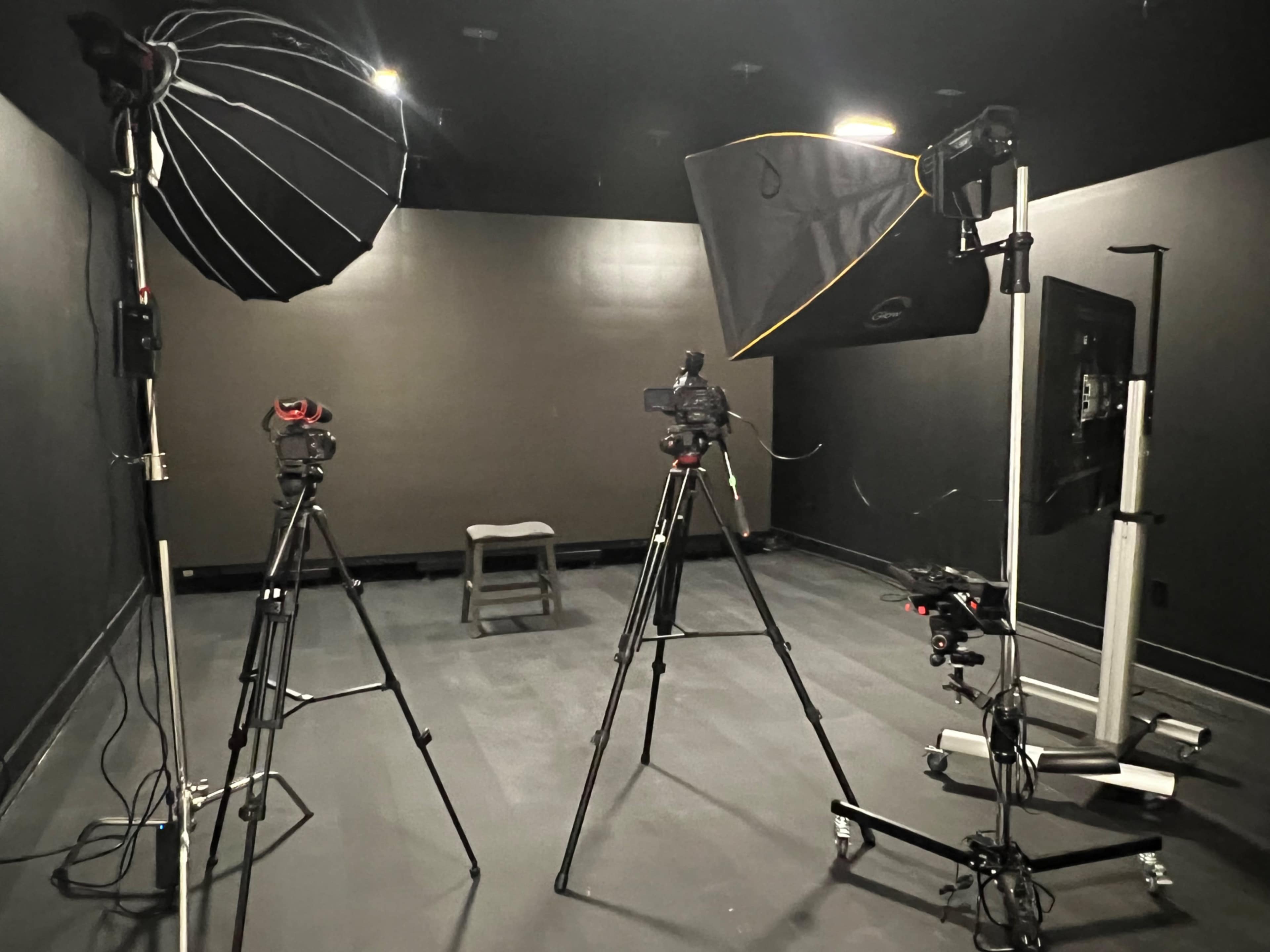 A photography studio equipped with two lighting setups and three cameras on tripods facing a single wooden stool in a dark, minimalist environment.