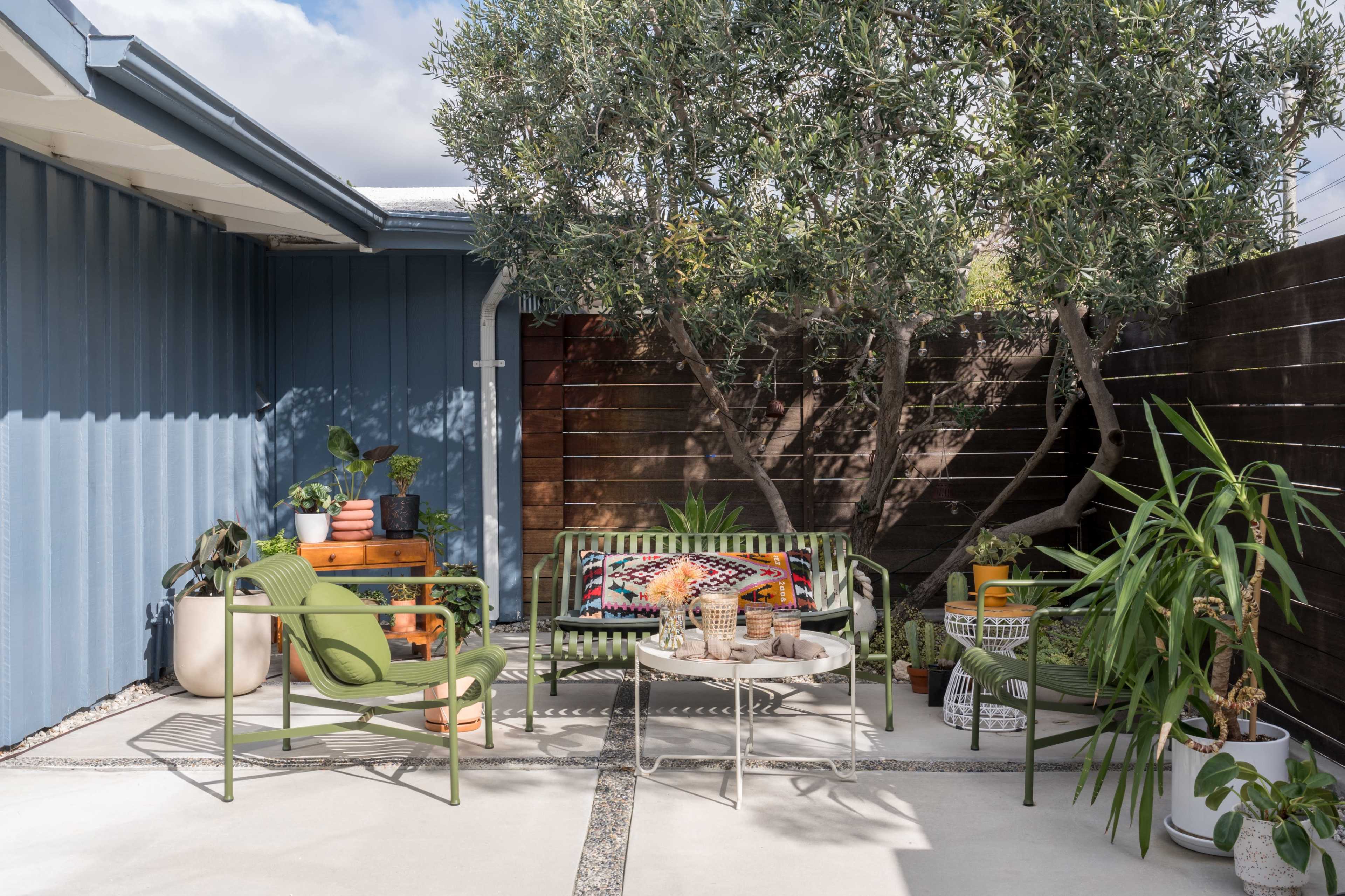 A cozy outdoor patio features a green metal bench, a table with various plants, and a large tree providing shade.